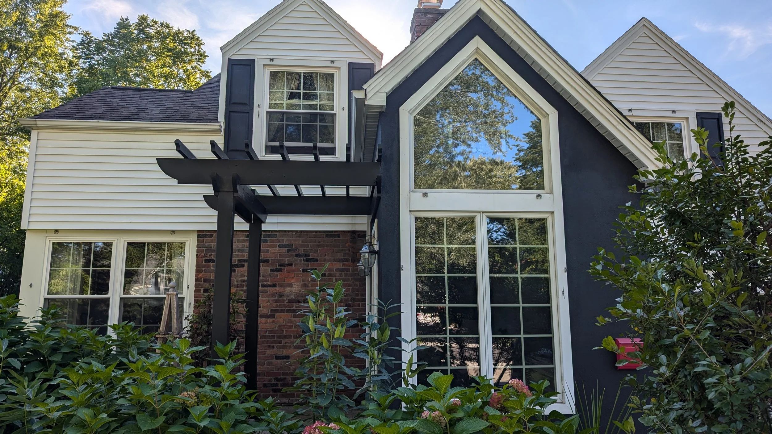 A house with large windows, vinyl siding, and a small brick section at the bottom. There is a black pergola with slats above the door, green plants and trees around the house, and a clear blue sky.