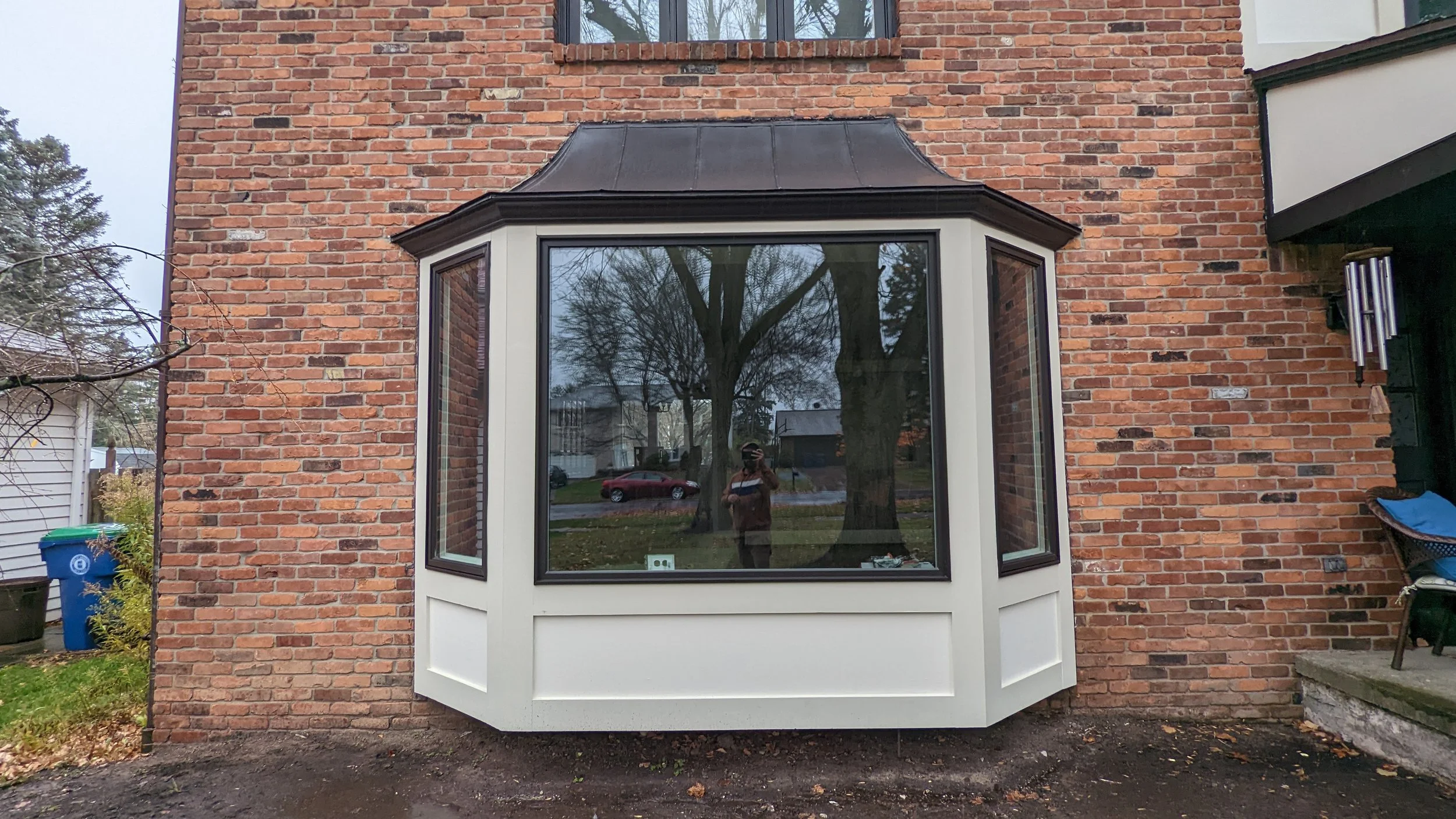 Back of a brick house with a large bay window that has white trim, reflecting trees and a person taking the photo. There are some trees and a street with parked cars in the background.