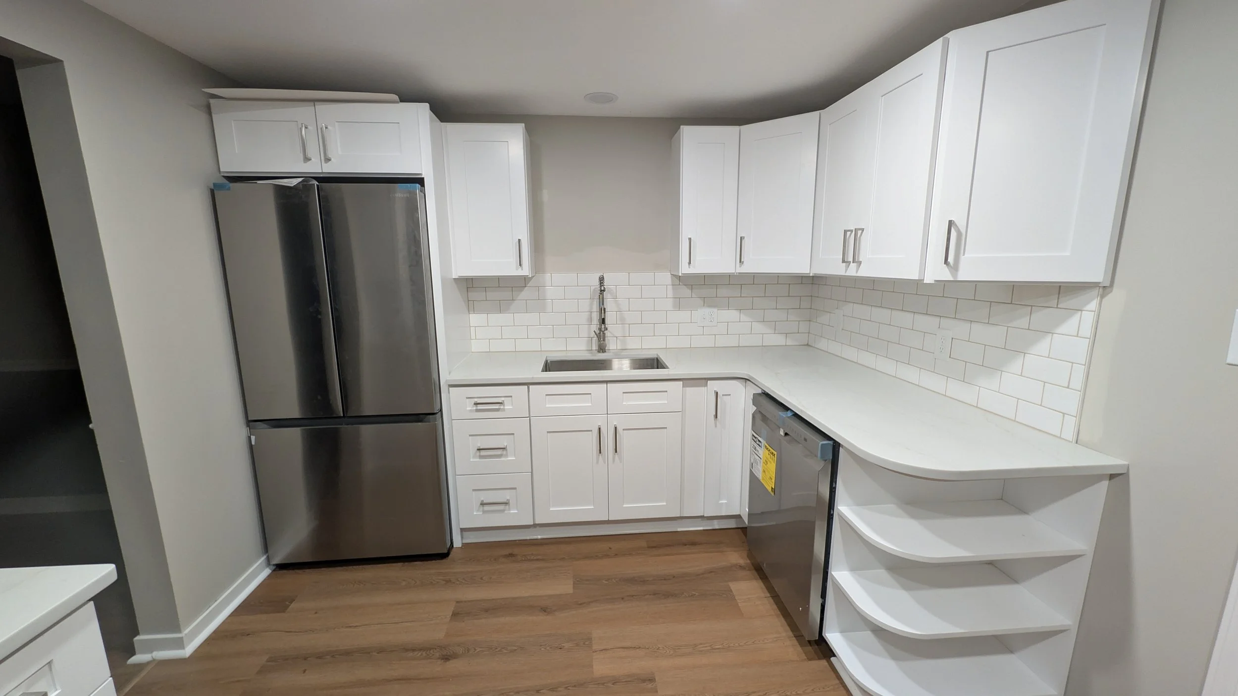 Modern kitchen with white cabinets, stainless steel refrigerator, white subway tile backsplash, white countertop, and open shelving on the right side.