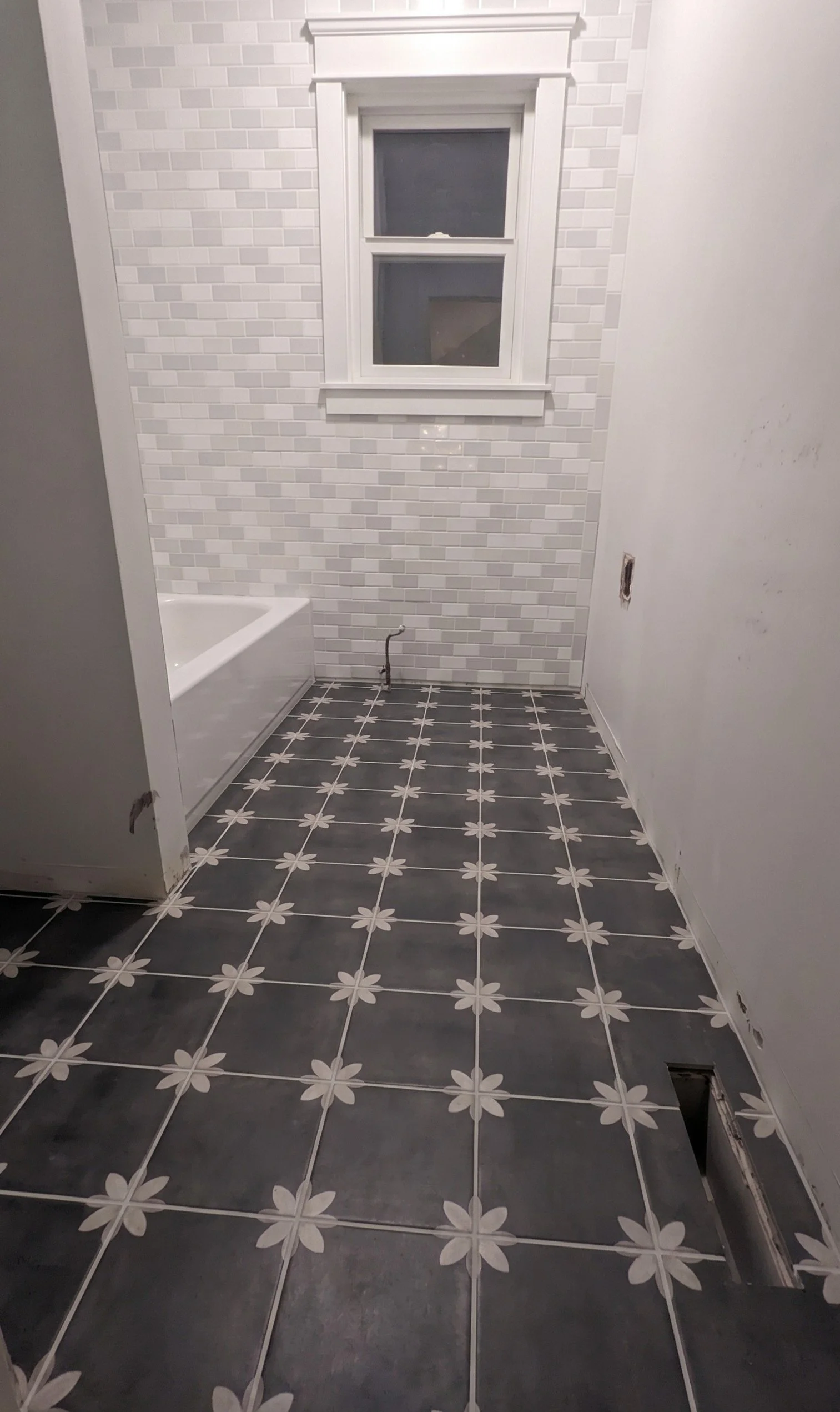 Empty bathroom with black and white floral patterned floor tiles, white subway tile walls, a small window, and an unfinished wall with holes.