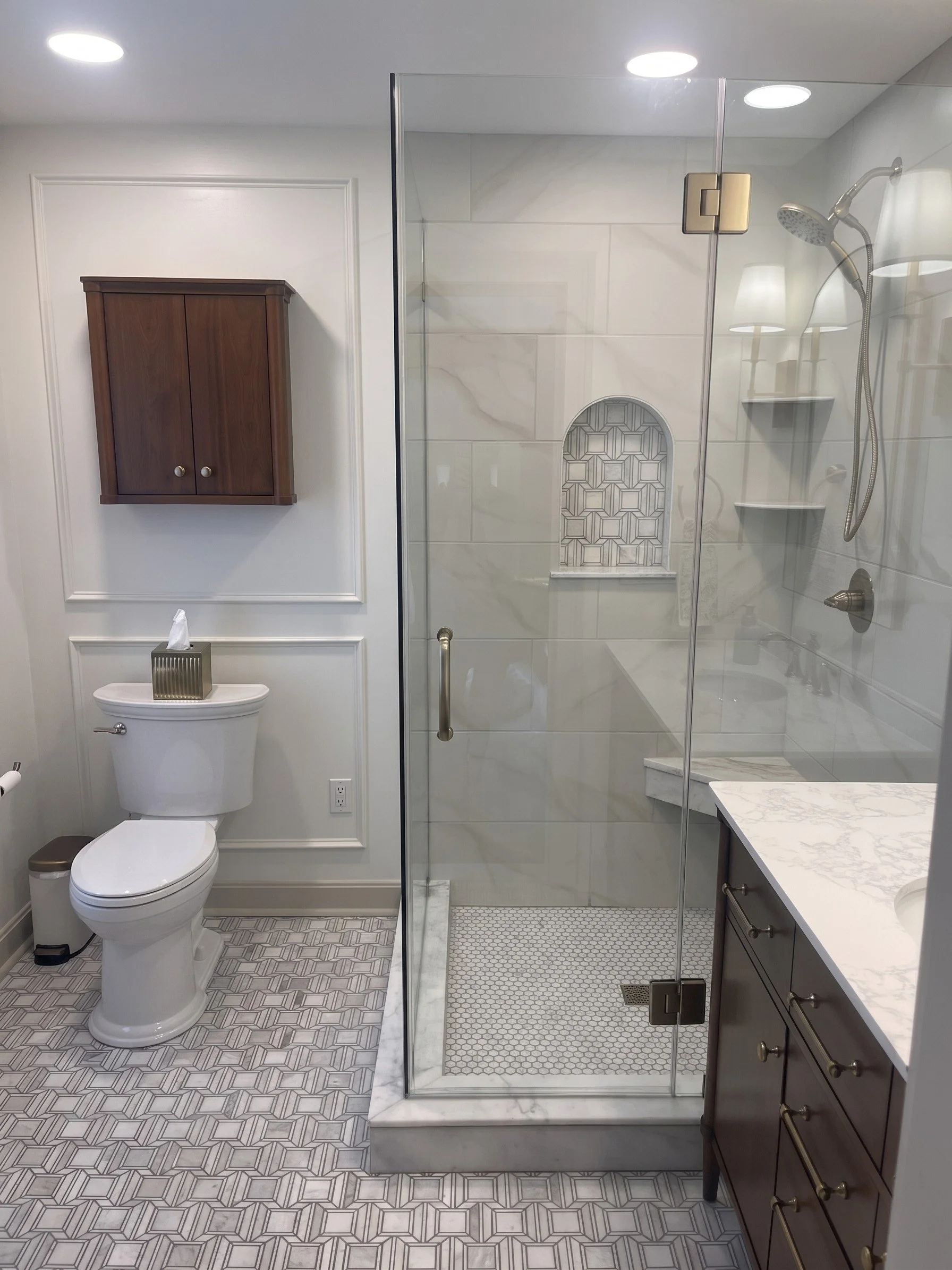 A modern bathroom with a toilet, wall-mounted wooden cabinet, glass shower enclosure, marble slab on the vanity, patterned floor tiles, and wall with white paneling and molding.