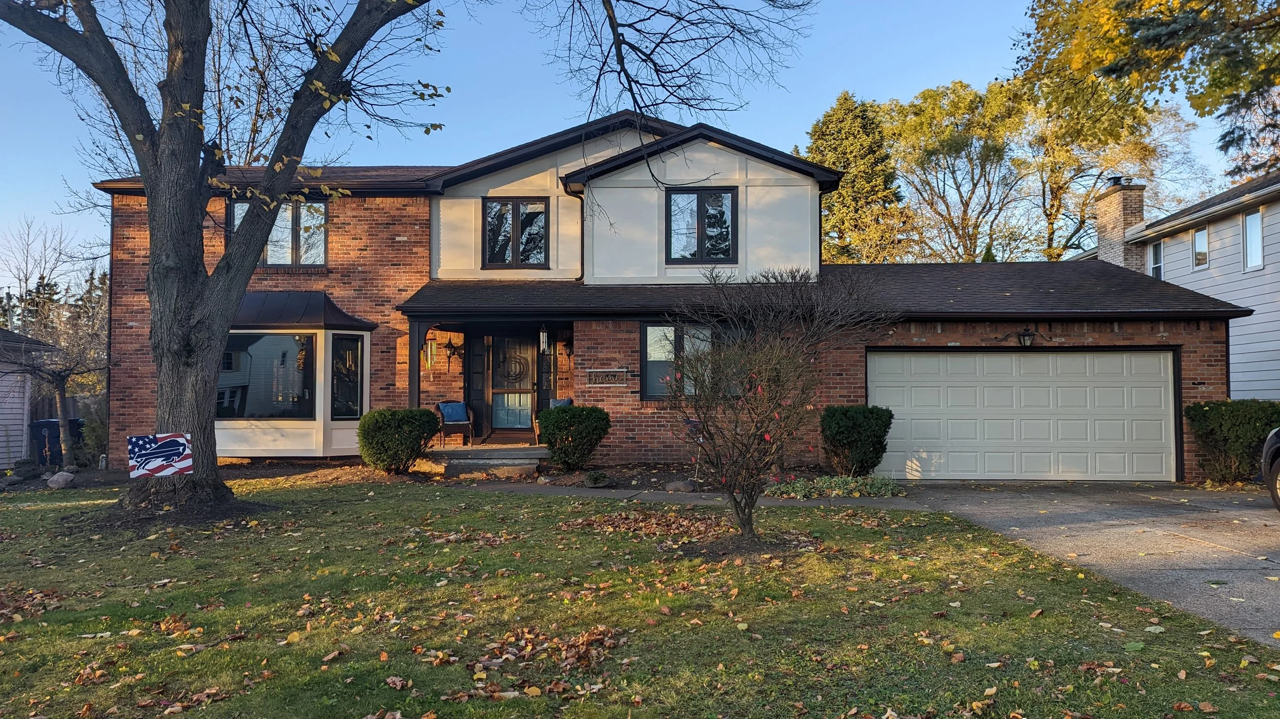 A two-story brick house with a tree in front and an American flag sign on the lawn. The house has a white and brick exterior, black window frames, and a two-car garage. The front yard has a lawn with fallen leaves.