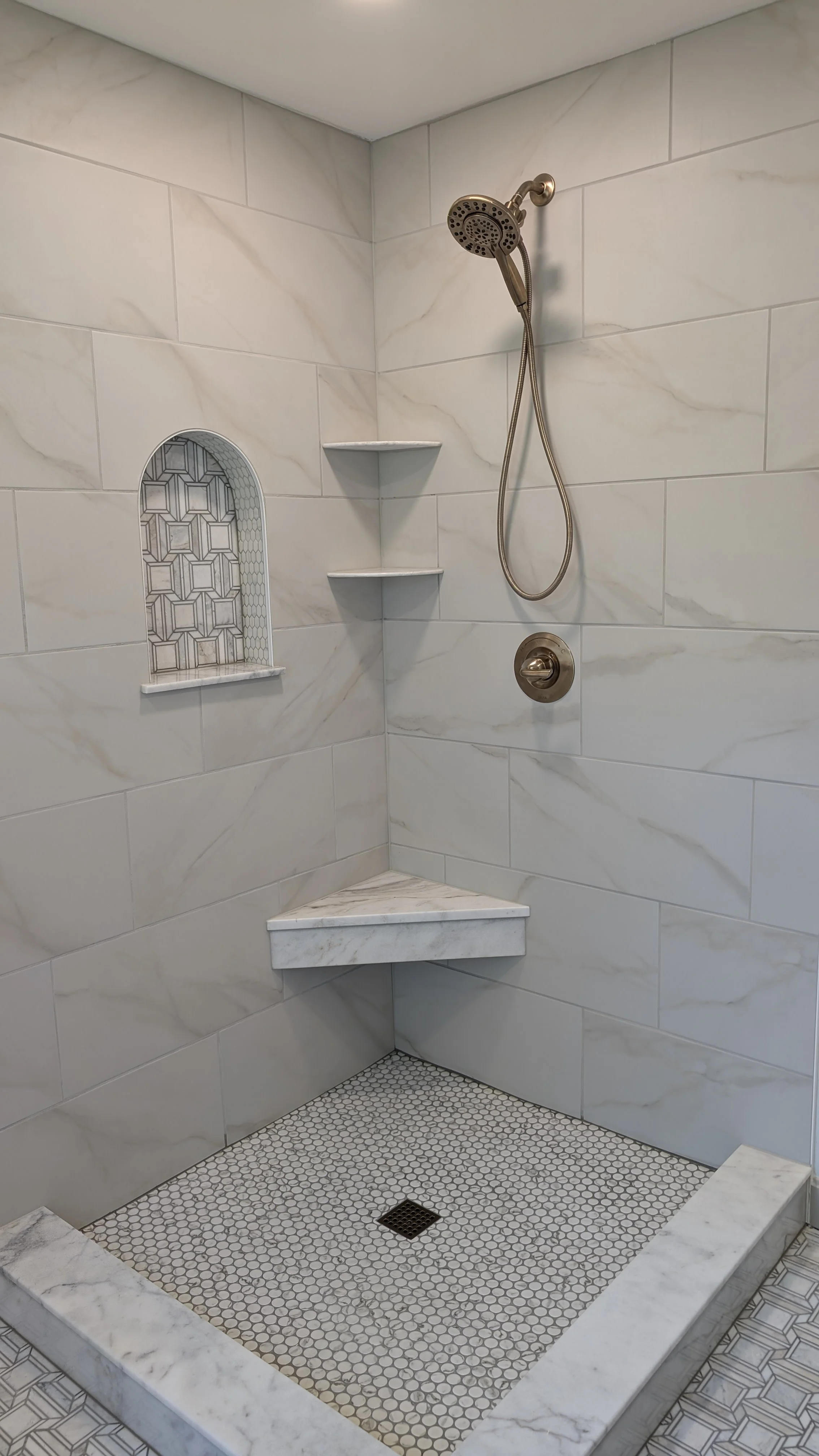 A walk-in shower with white marble tiled walls, a built-in niche with patterned tiles, two corner shelves, a marble bench, and small white hexagon shower floor tiles with a drain in the center.