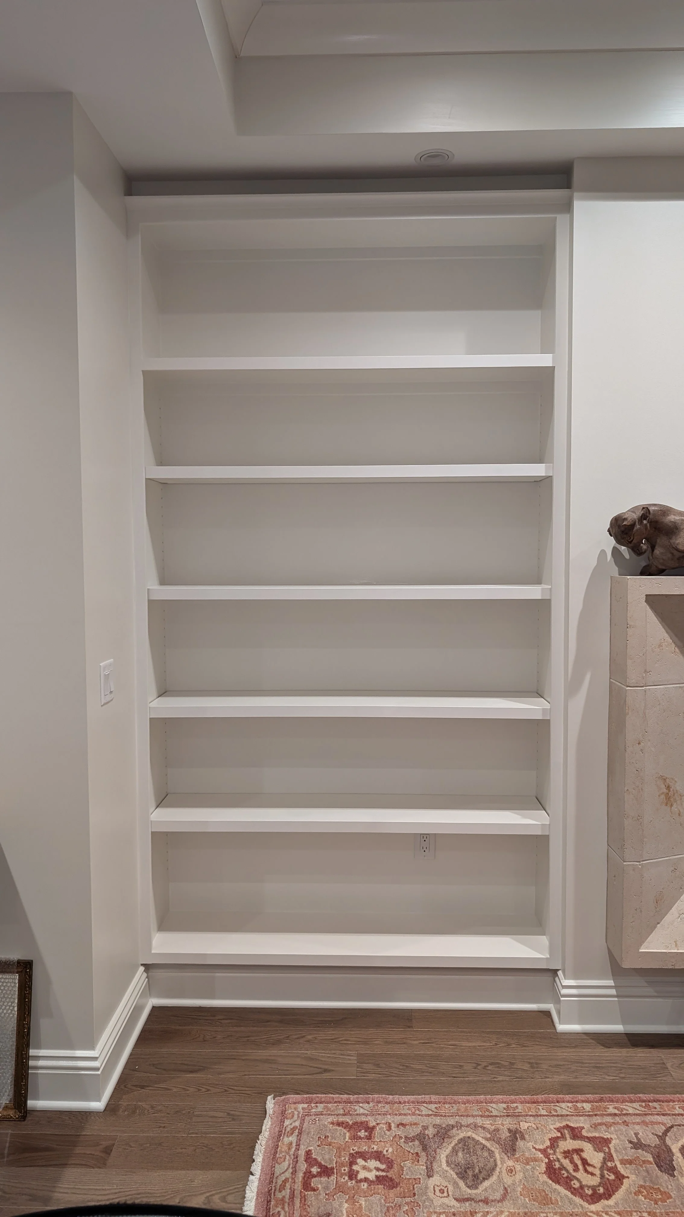 Empty white built-in bookshelf with eight shelves, located in a room with wood flooring, a pink and beige rug in the foreground, and a small sculpture on a nearby counter to the right.