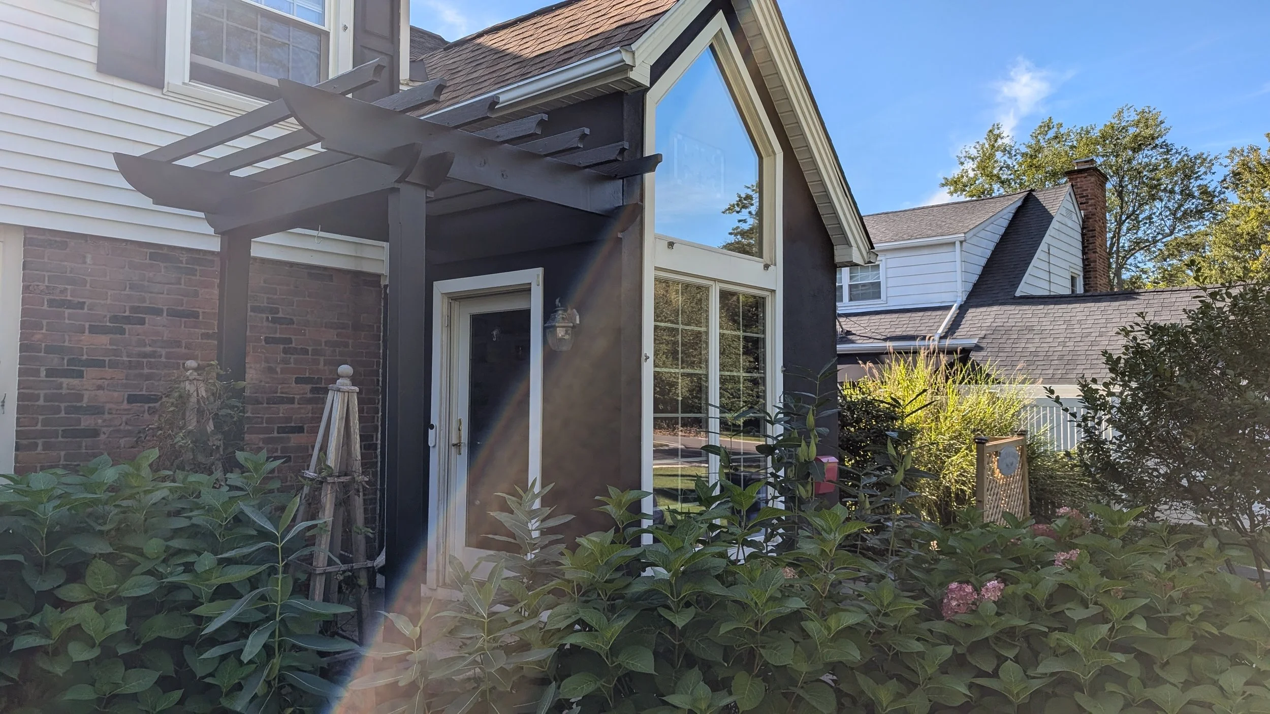 Backyard with a house featuring a brick and siding exterior, large windows, and a small porch with a white door. There are green plants and bushes in the yard.