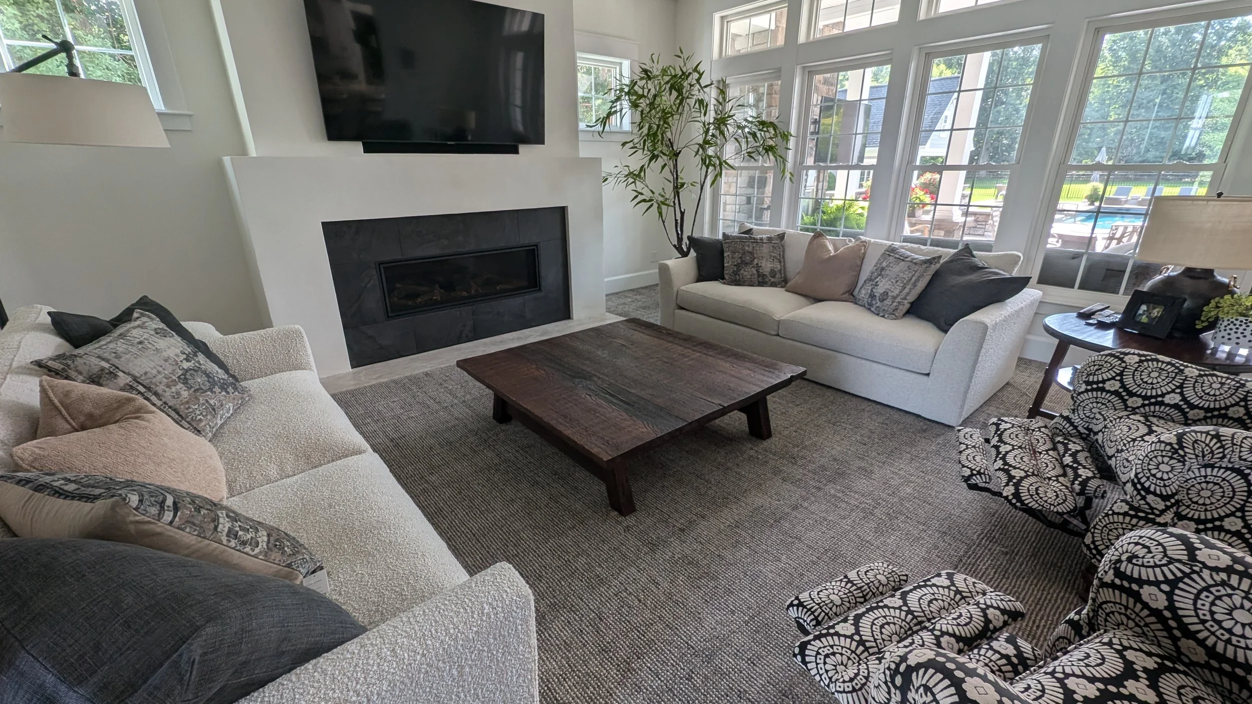 A cozy living room with large windows, a fireplace, a wall-mounted TV, and multiple sofas with assorted pillows, a wooden coffee table, and a side table with a lamp and photo frames. Outside view shows a backyard with a pool and patio.