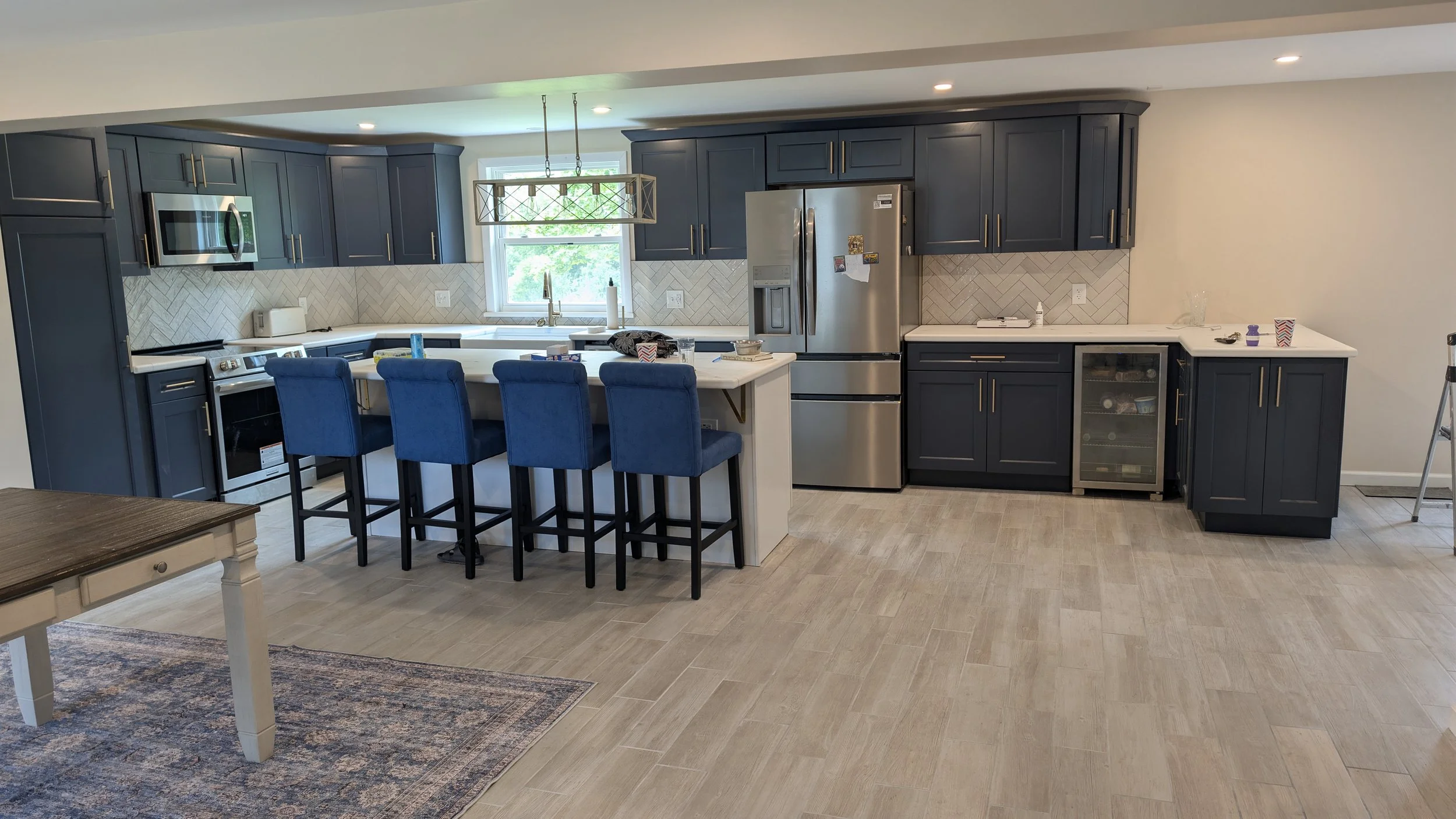 Modern kitchen with dark blue cabinets, stainless steel appliances, and a white countertop island with four blue chairs. There is a window above the sink, and a light fixture hangs over the island. The floor is light wood, and there is a rug partiall