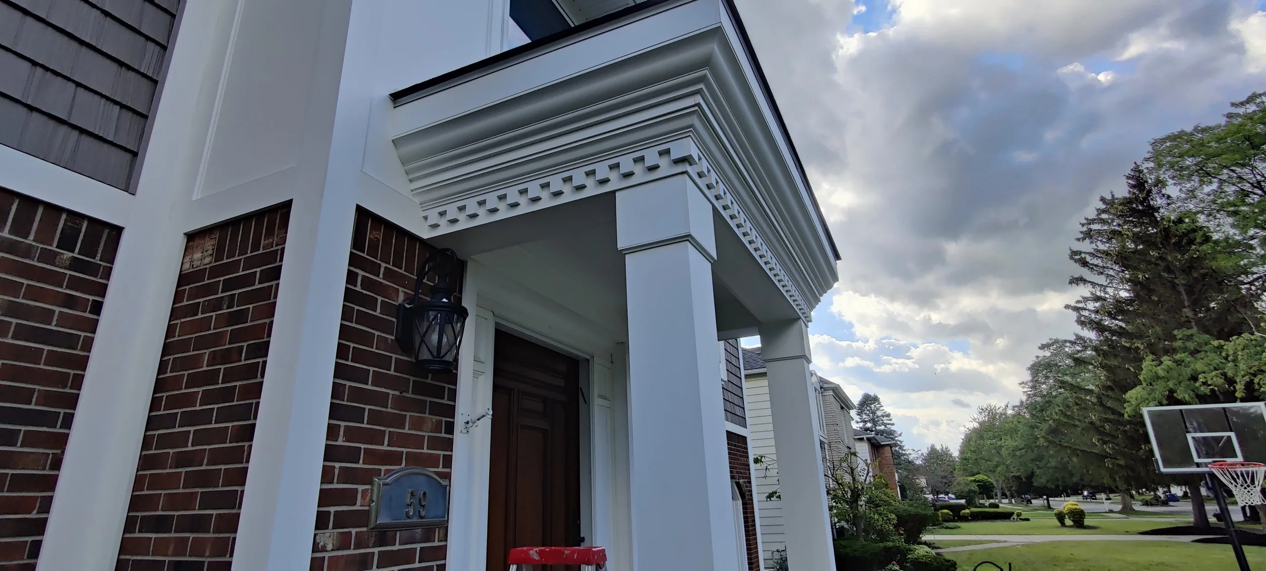 Close-up of a house's entrance with a brick wall, white columns, and a decorative white corbel under the overhang, with a black lantern light fixture and house number 55. The sky is partly cloudy, and a basketball hoop is visible in the background in