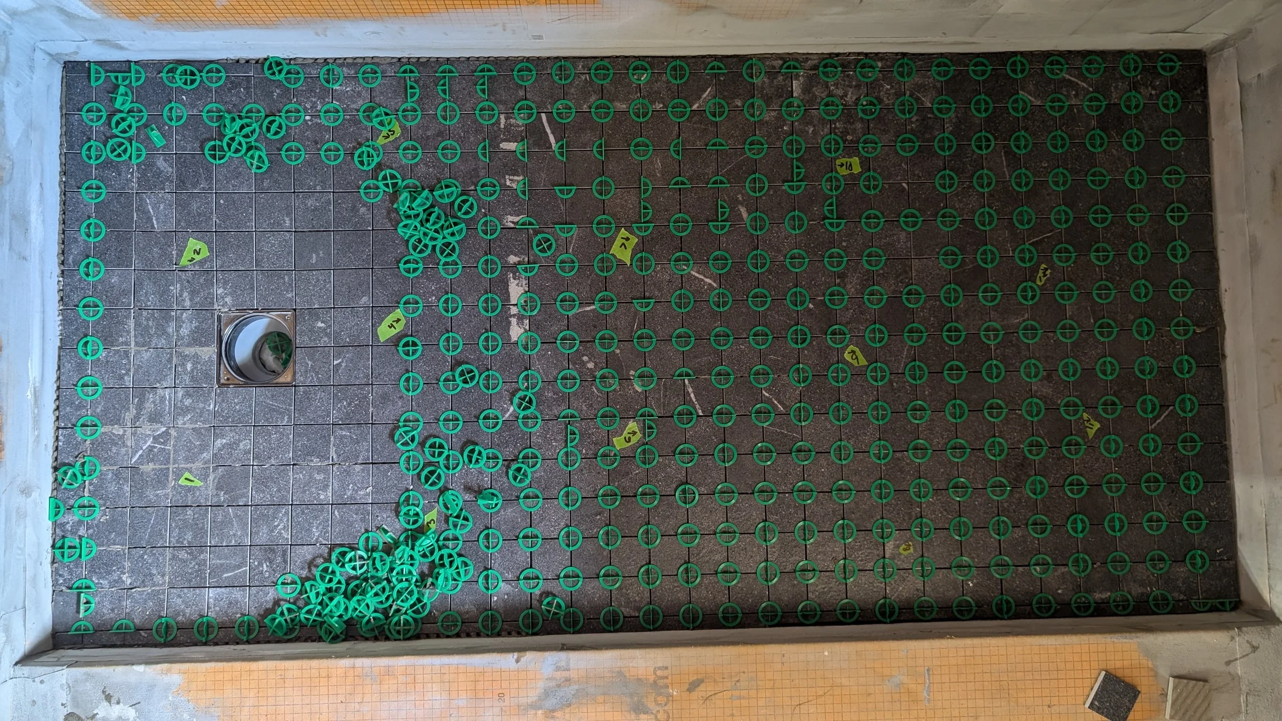 A tiled floor under construction with numerous small green plastic caps arranged in a pattern and some scattered on the floor, along with several yellow markers with numbers. There is a drainage grate in the lower left corner.