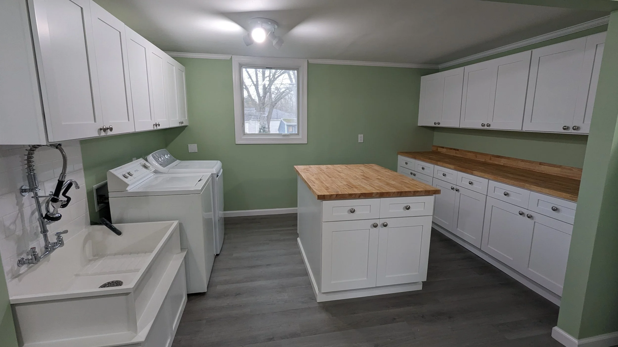 A laundry room with white cabinets, a window showing a tree outside, a washer and dryer, a utility sink, and wooden countertops, painted light green.