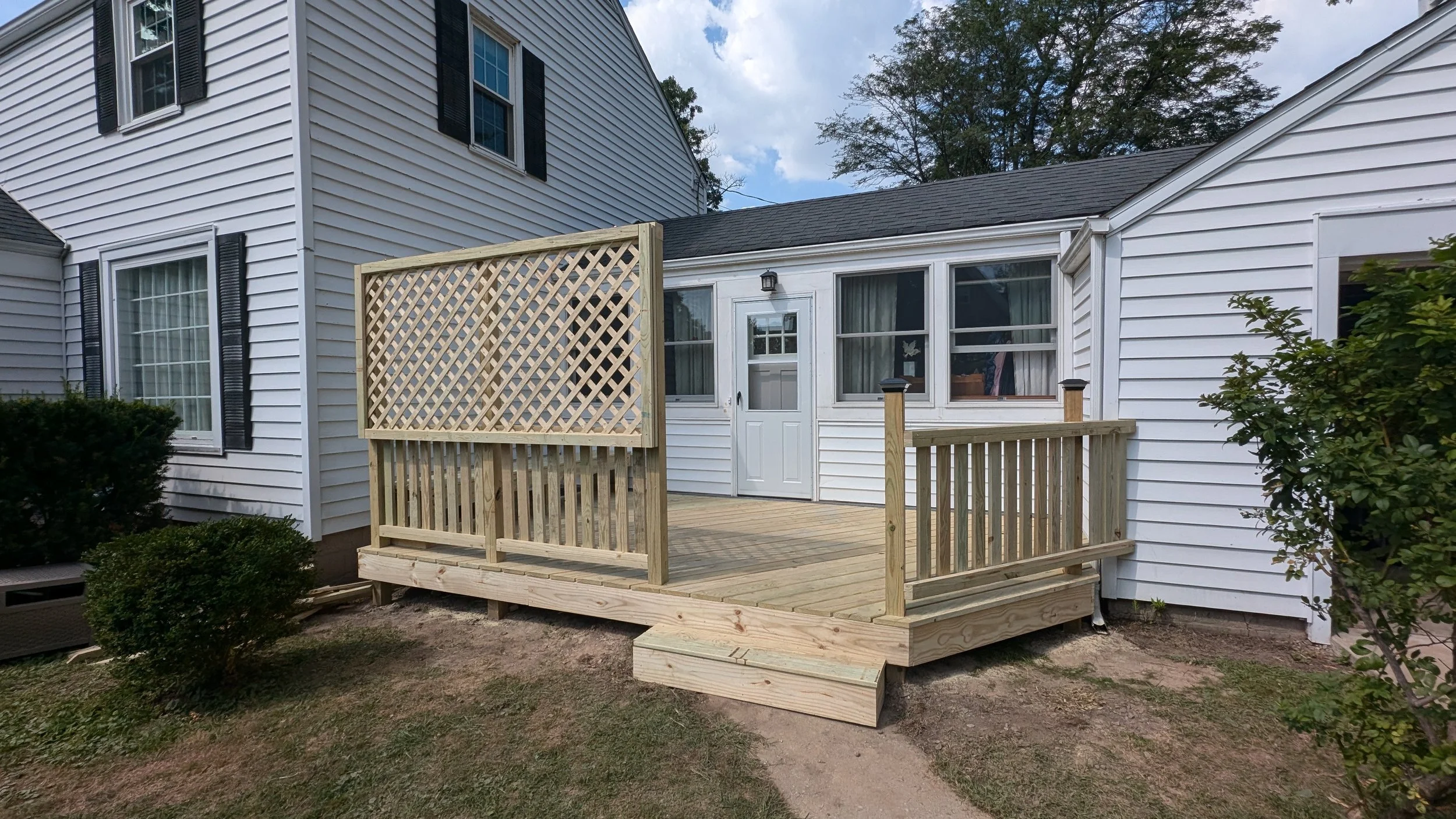 Backyard wooden deck under construction attached to a white house with black shutters, trees, and partly cloudy sky in the background.