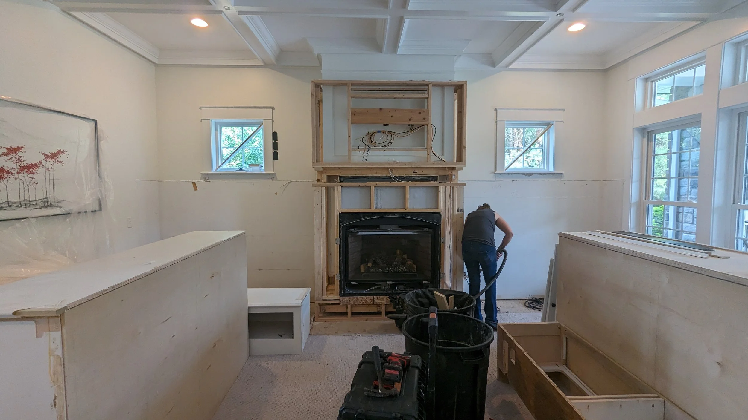 A person working on home renovation in a living room, installing a built-in fireplace surrounded by unfinished wooden framing, with windows and a painting on the wall.