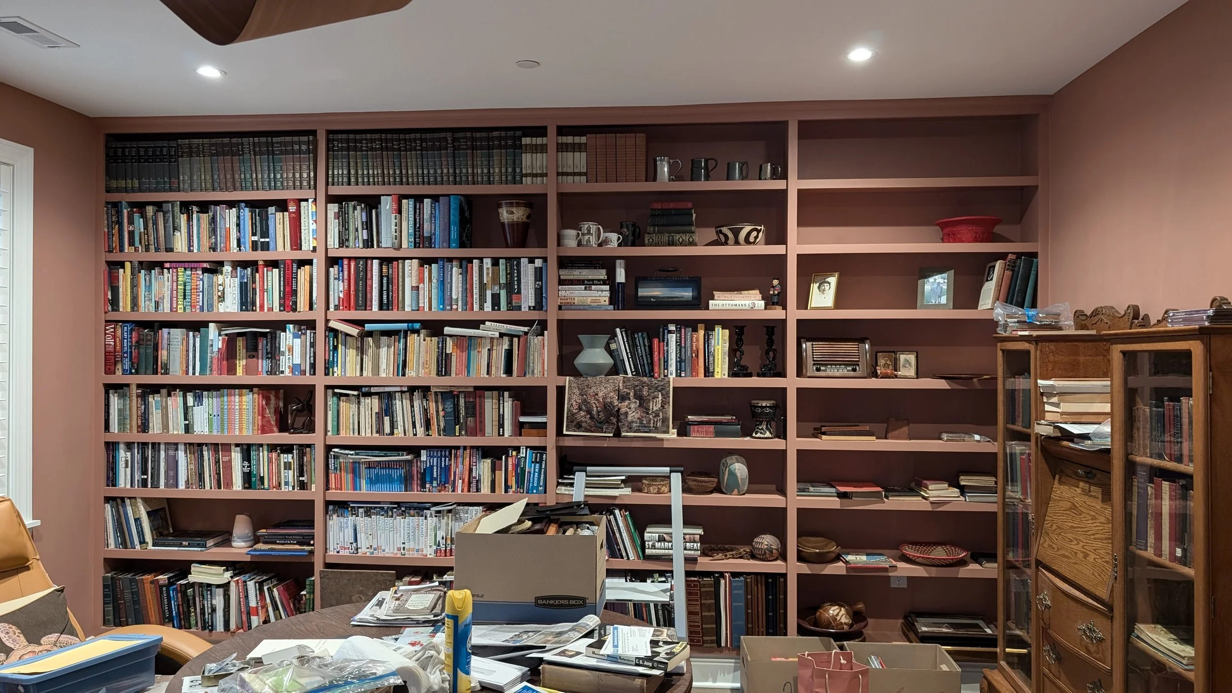 A large bookshelf filled with books and decorative items against a pink wall in a cluttered room.