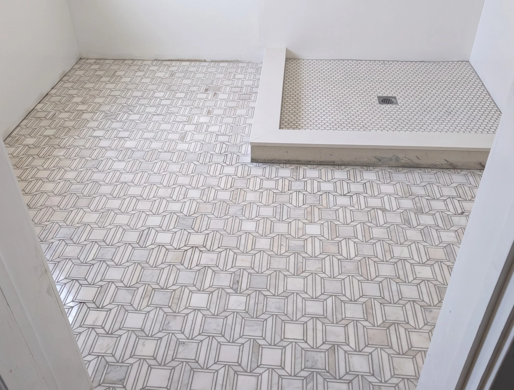 A bathroom floor with geometric patterned tiles and a shower area with small hexagonal tiles and a drain in the center.