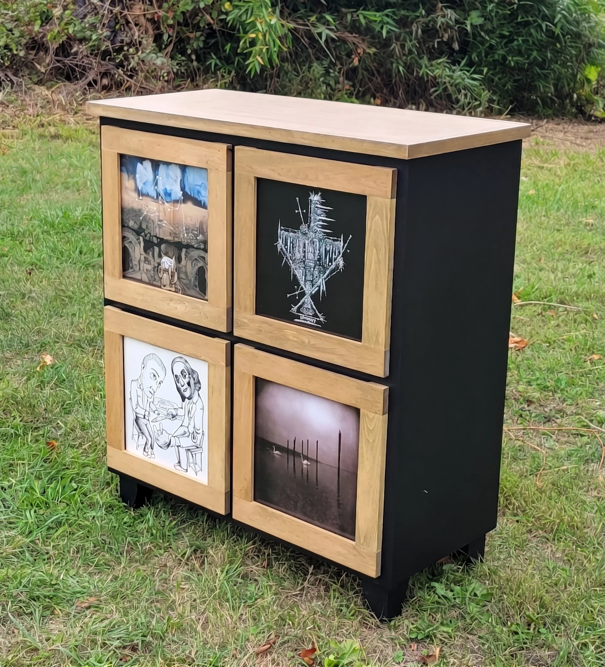 A small cabinet with a wooden frame and black sides, displaying various posters and drawings on its doors, located outdoors on grass with bushes in the background.
