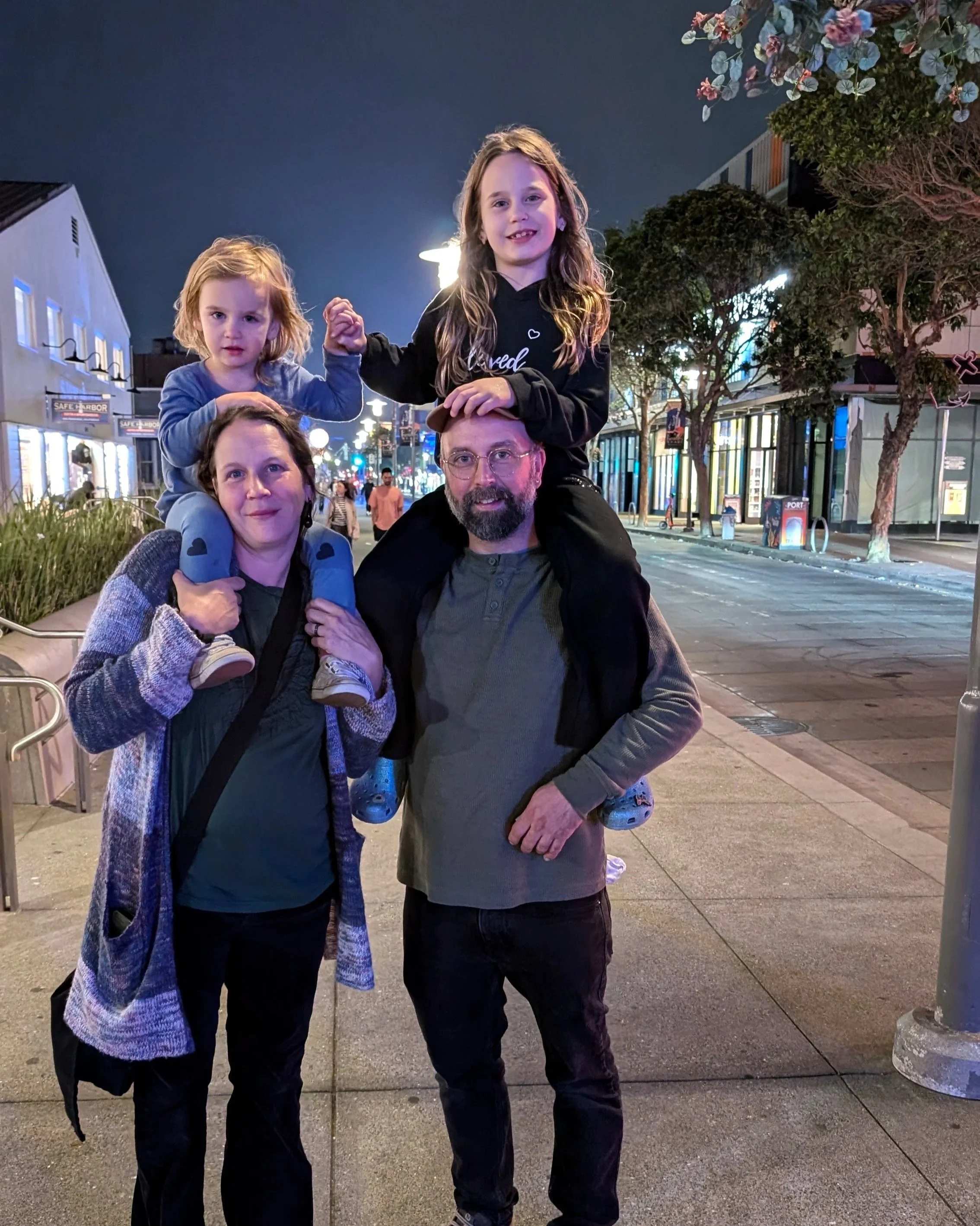 A family of four with two young girls, a woman, and a man, posing together on a city street at night. The girls are sitting on the shoulders of the adults, holding hands and smiling, with trees and lit buildings in the background.