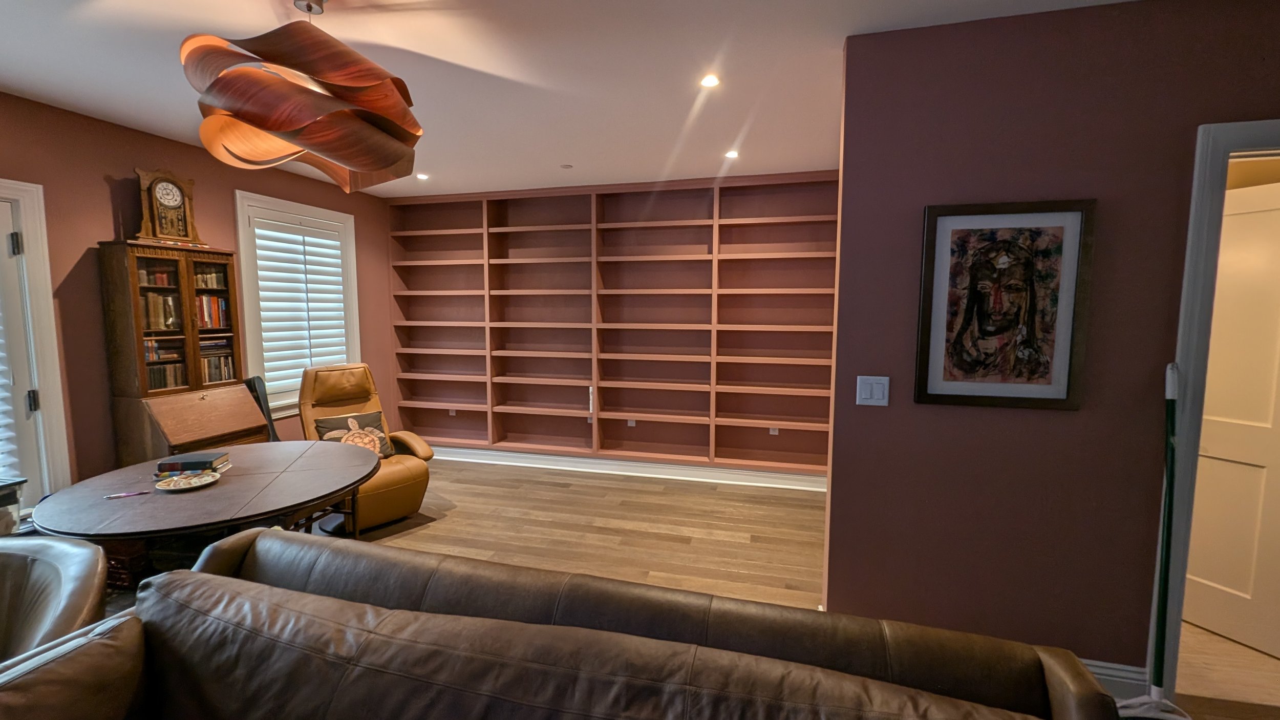 A living room with a large, empty wooden bookshelf, a round dining table with chairs, a leather armchair, a framed abstract art piece on the wall, and a wooden bookcase with books, all in warm lighting.