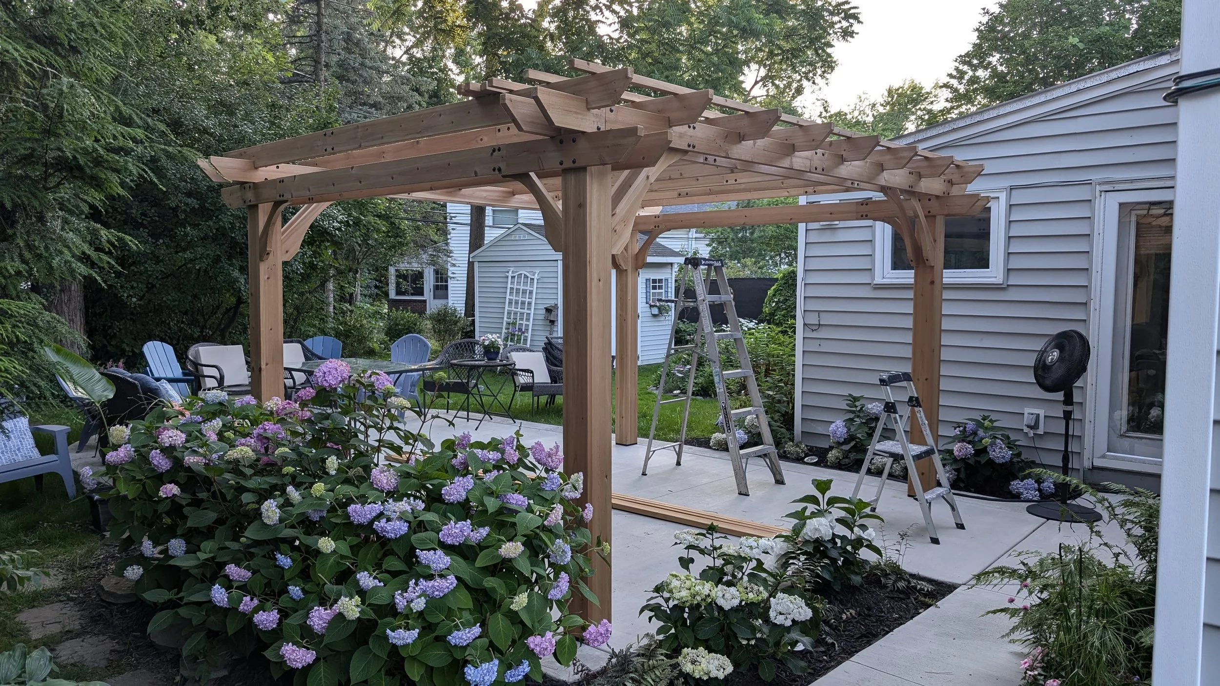 A backyard patio under construction with a wooden pergola frame, surrounded by flowering plants and outdoor furniture.