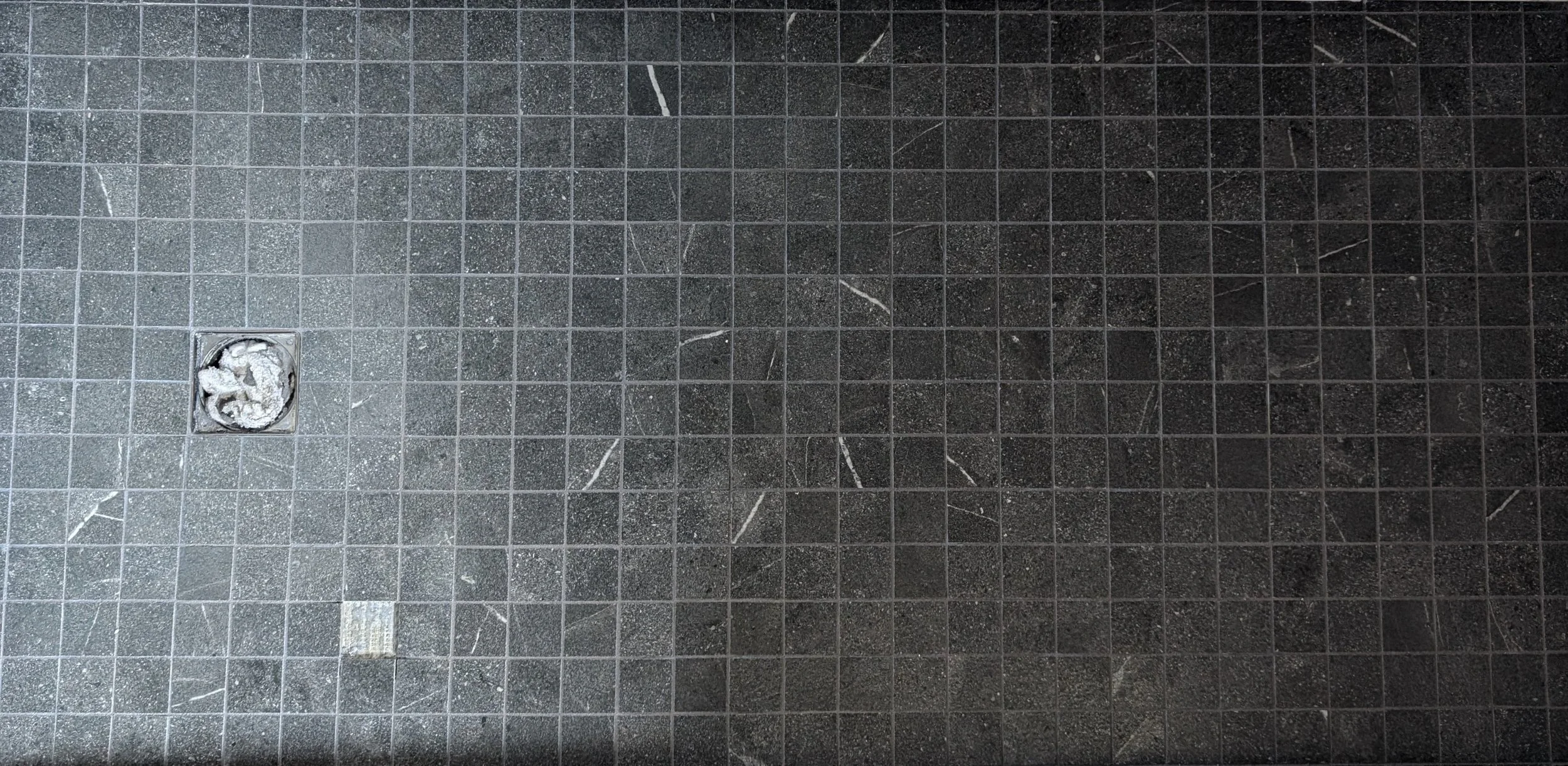 A dark gray tiled bathroom or shower floor with a square drain in the lower left corner. The drain cover is removed, revealing debris inside.