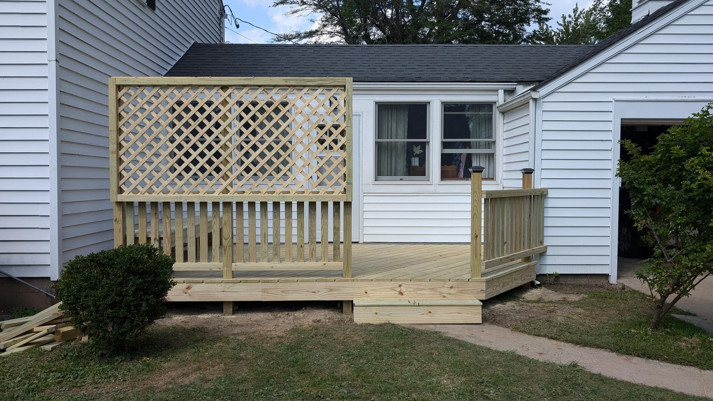 New wooden deck with a lattice privacy screen attached to the back of a white house, with some bushes and green grass in the yard.