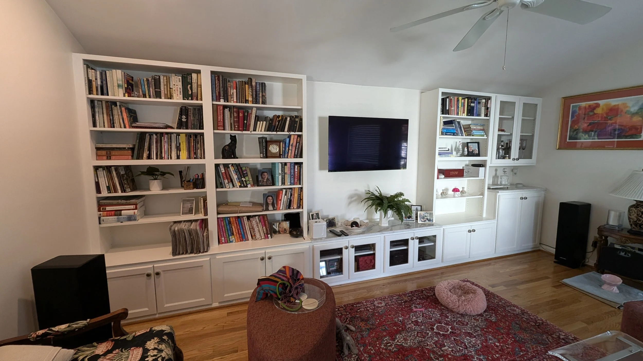 Living room with white built-in bookshelves, a mounted flat-screen TV, a potted plant, framed photographs, and a red patterned rug.