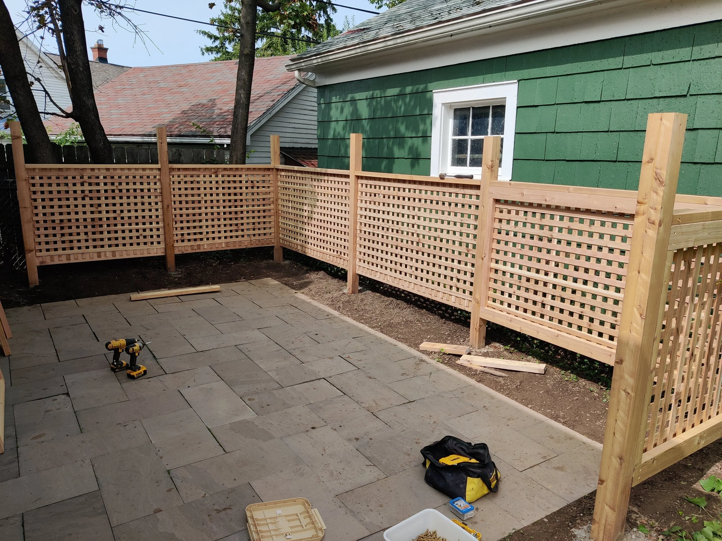 A backyard with a partially built wooden privacy fence. Pavers are laid on the ground, and there are tools and construction materials on the patio. A green house with white window trim is in the background, along with trees and neighboring houses.