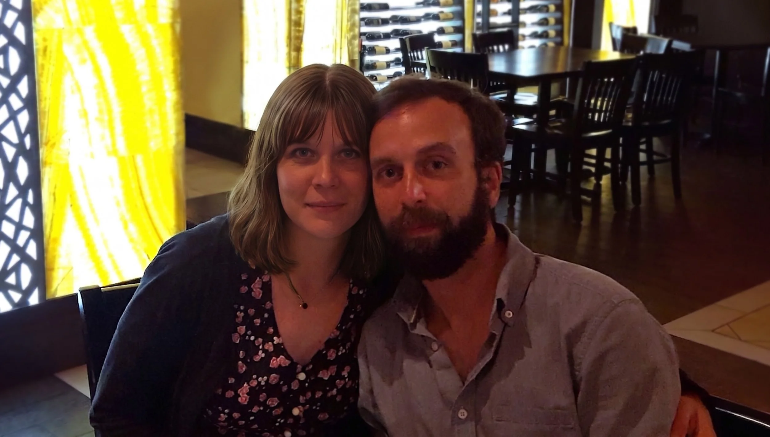 A couple taking a selfie in a restaurant with yellow curtains and wine bottles in the background.