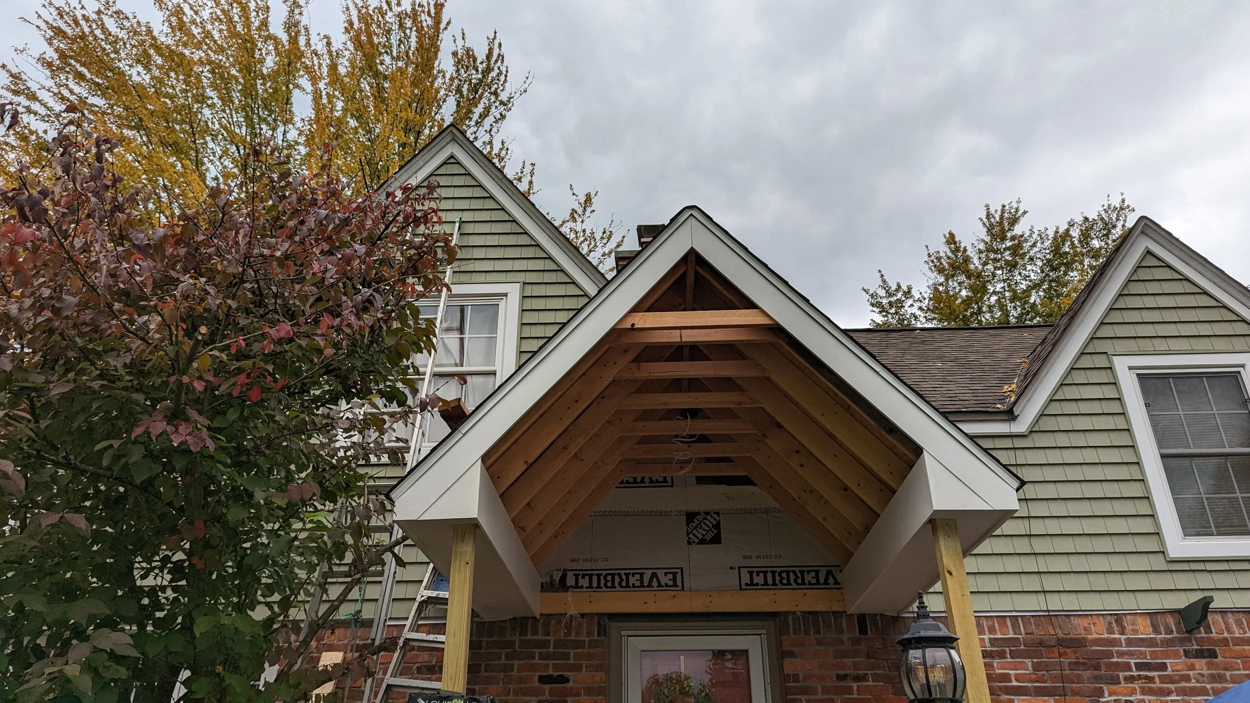 The image shows an unfinished porch extension under construction on a house with green siding. The porch has a wooden frame with an overhanging roof. A ladder is leaning against the house, and there's a tree with reddish leaves on the left and trees 
