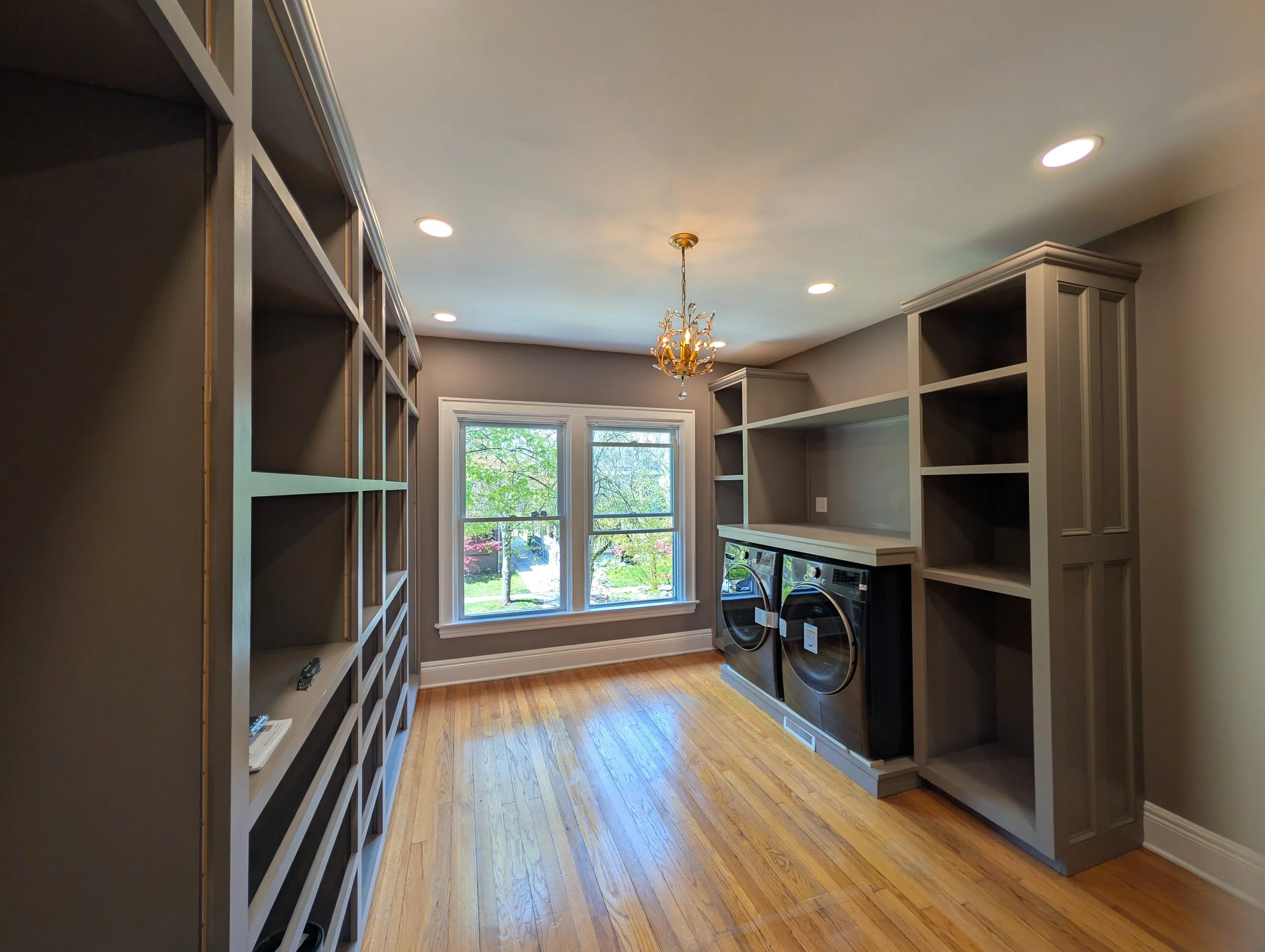 A laundry room with a large window showing trees outside, parquet wood flooring, built-in white shelves, a chandelier, and a washer and dryer.