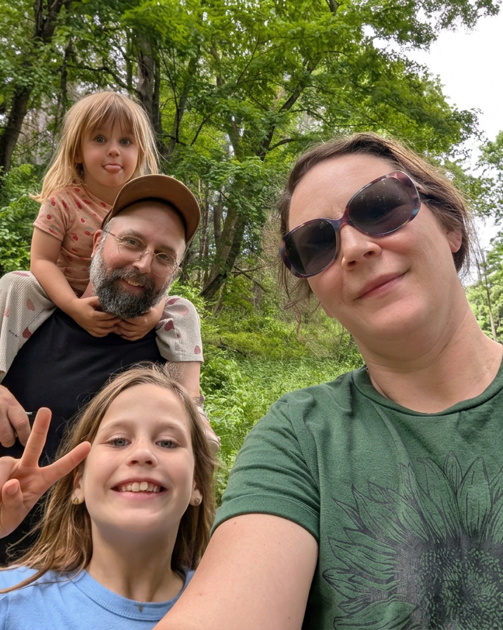 A woman and three children outdoors in a wooded area, taking a selfie. The woman wears large sunglasses and a green shirt, the girl in front has a peace sign and a big smile, the girl on man's shoulders has her tongue out, and the man has a beard, glasses, and a cap.