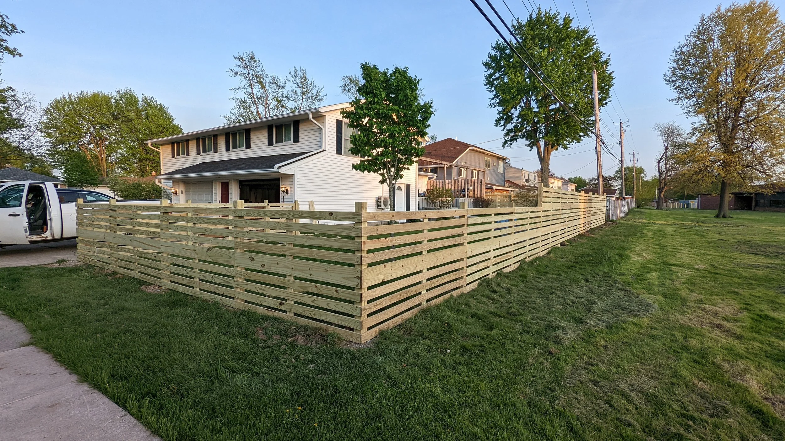 A residential backyard with a newly built wooden fence, green lawn, trees, and a white house with black shutters, a garage, and a pickup truck parked outside.