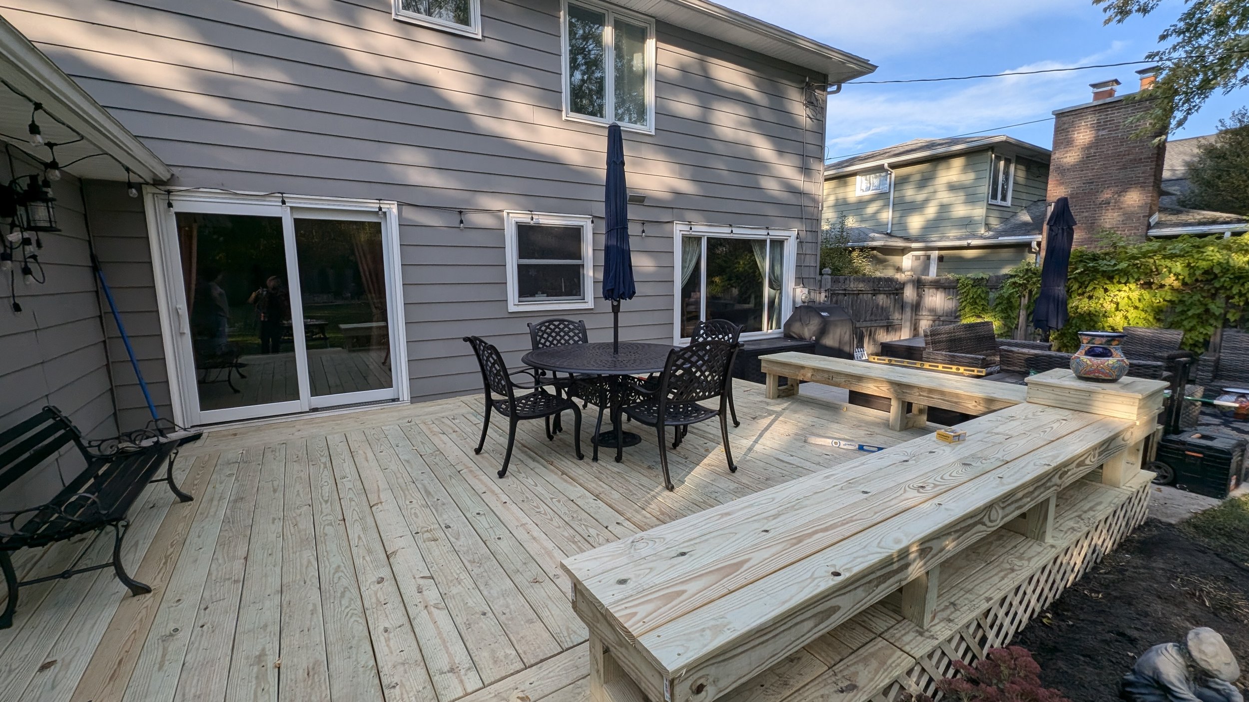 Backyard patio with new wooden decking, black metal dining table with chairs, blue patio umbrellas, outdoor grill, and outdoor furniture. House with gray siding and sliding glass door, surrounded by trees.