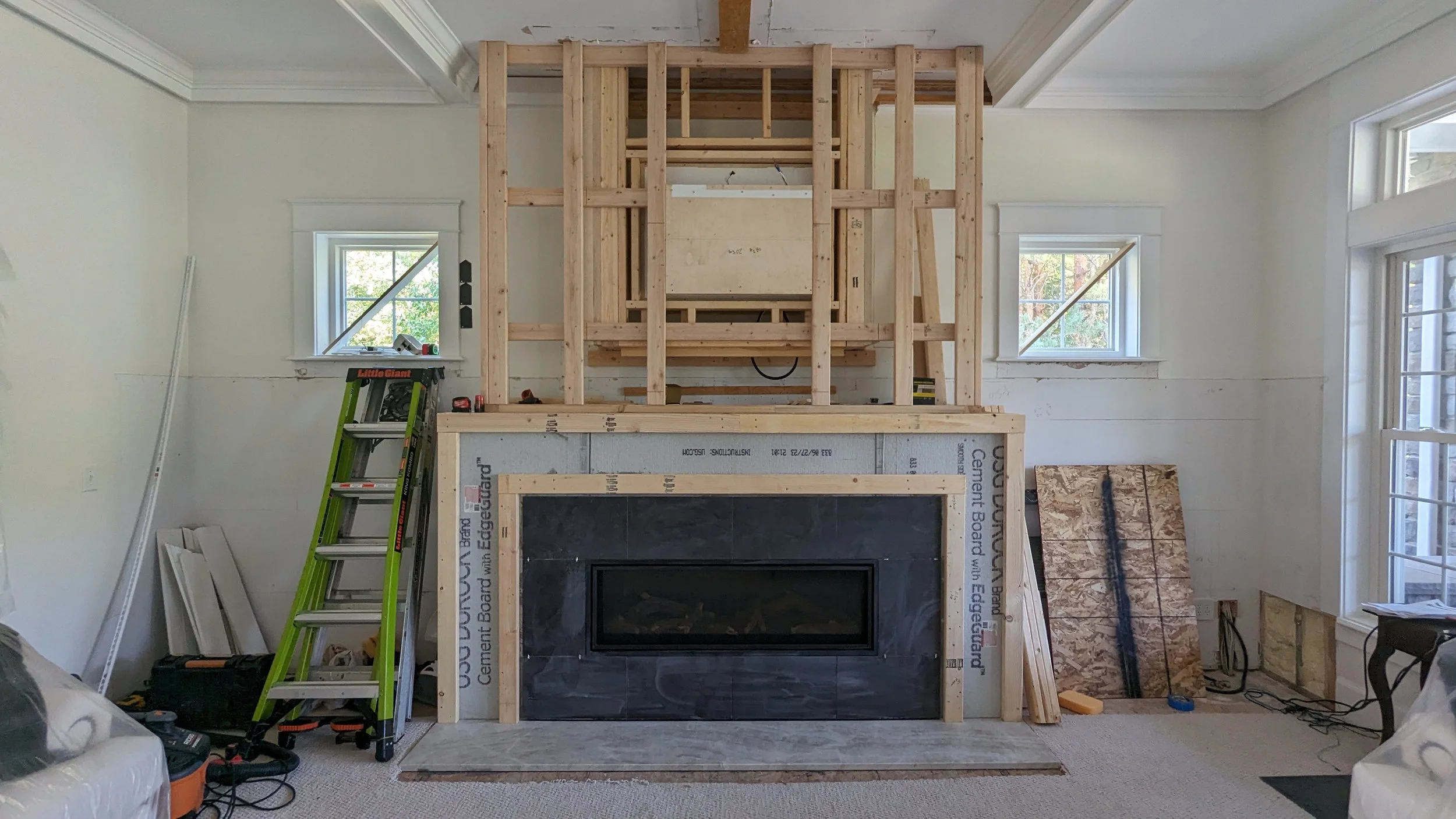 Inside a house under renovation, a fireplace with black surround is being installed with new wooden framing around it. A green ladder is leaning against the wall on the left, and construction tools and materials are scattered around.