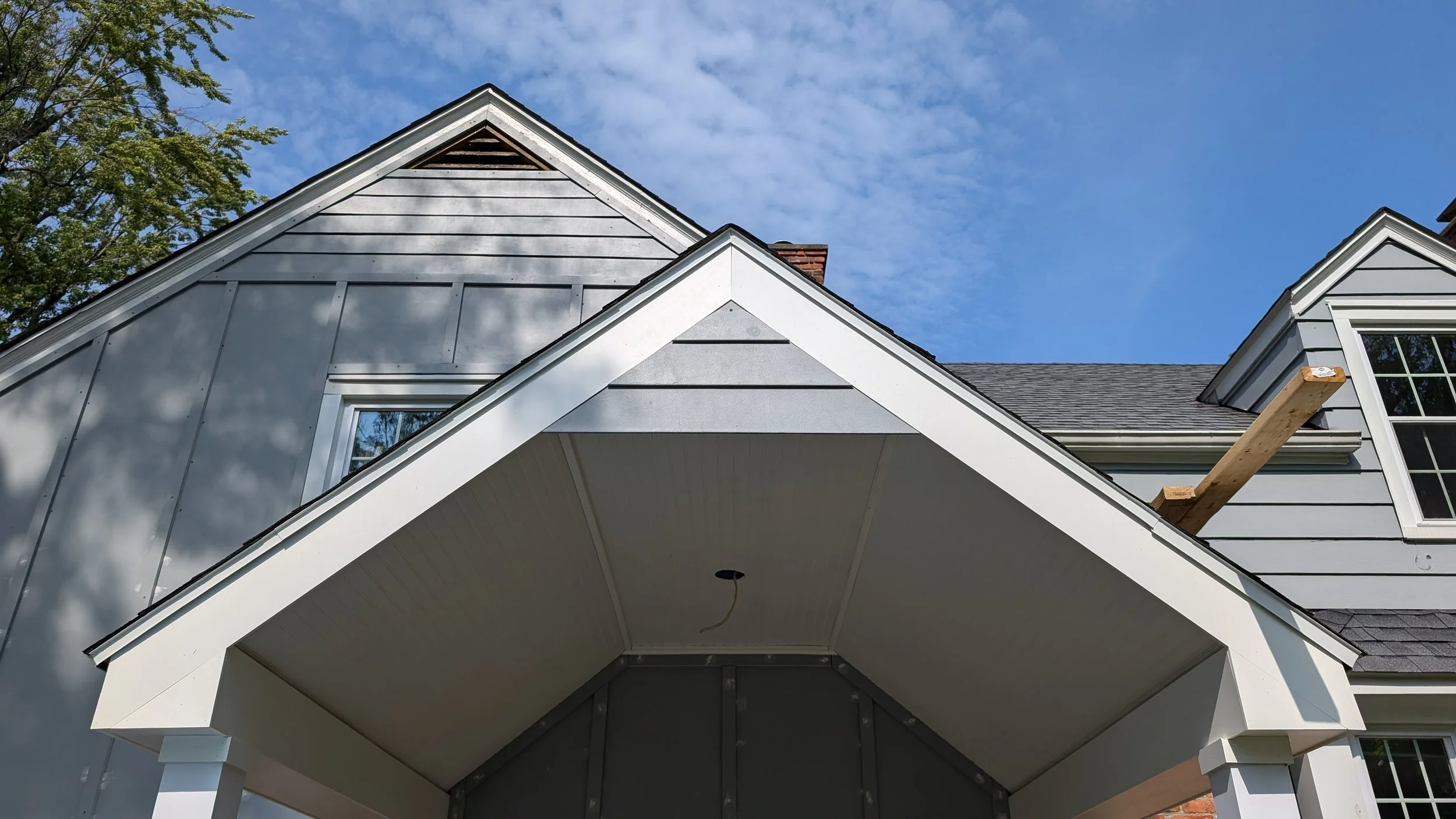 Close-up of a house's roof with new gray siding and trim, under a blue sky with some clouds and a tree in the background. A wooden board extends from the roof.