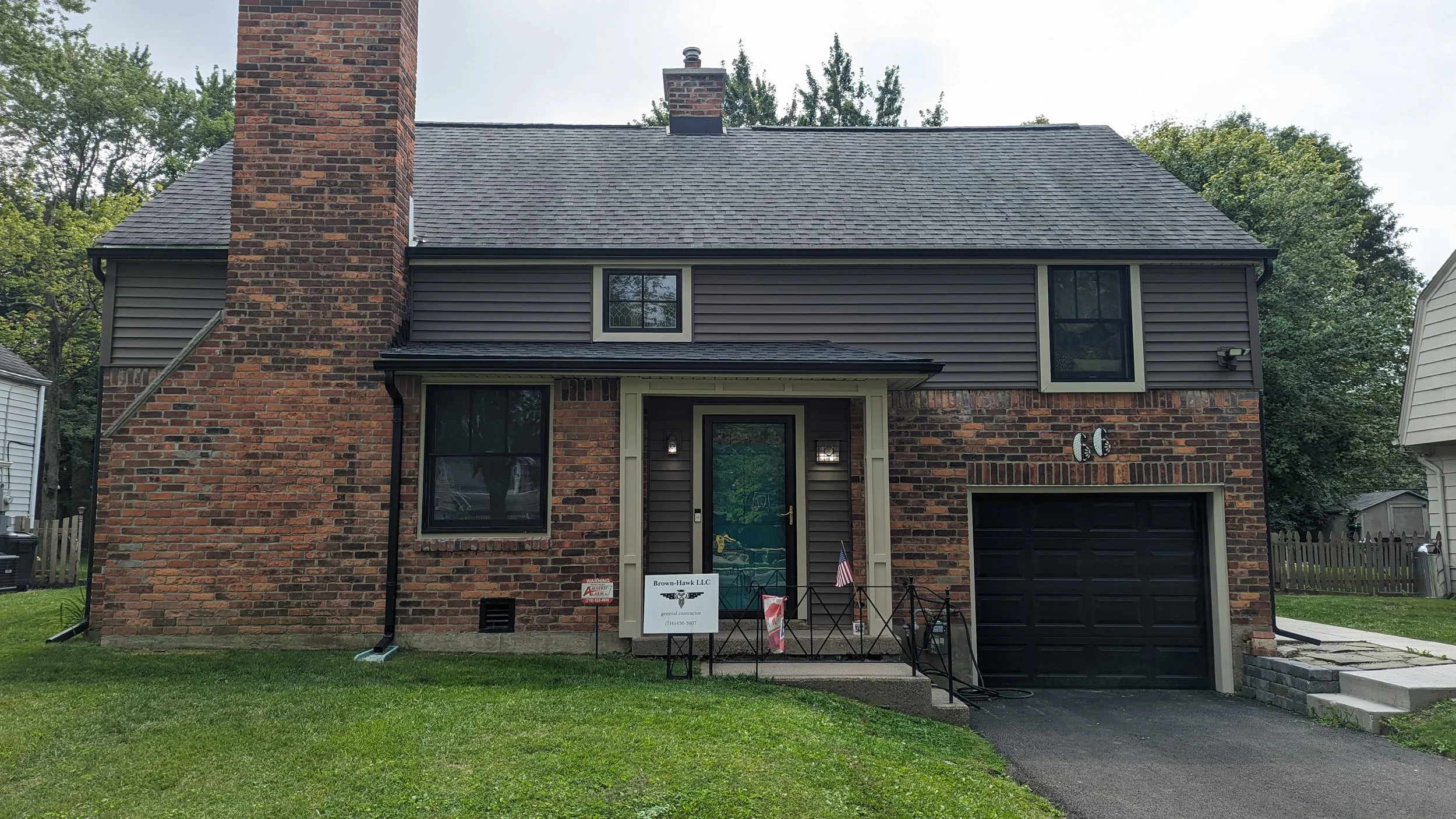 Front view of a brick and gray siding house with a black garage door, front door, and multiple windows, with a lawn and trees in the background.