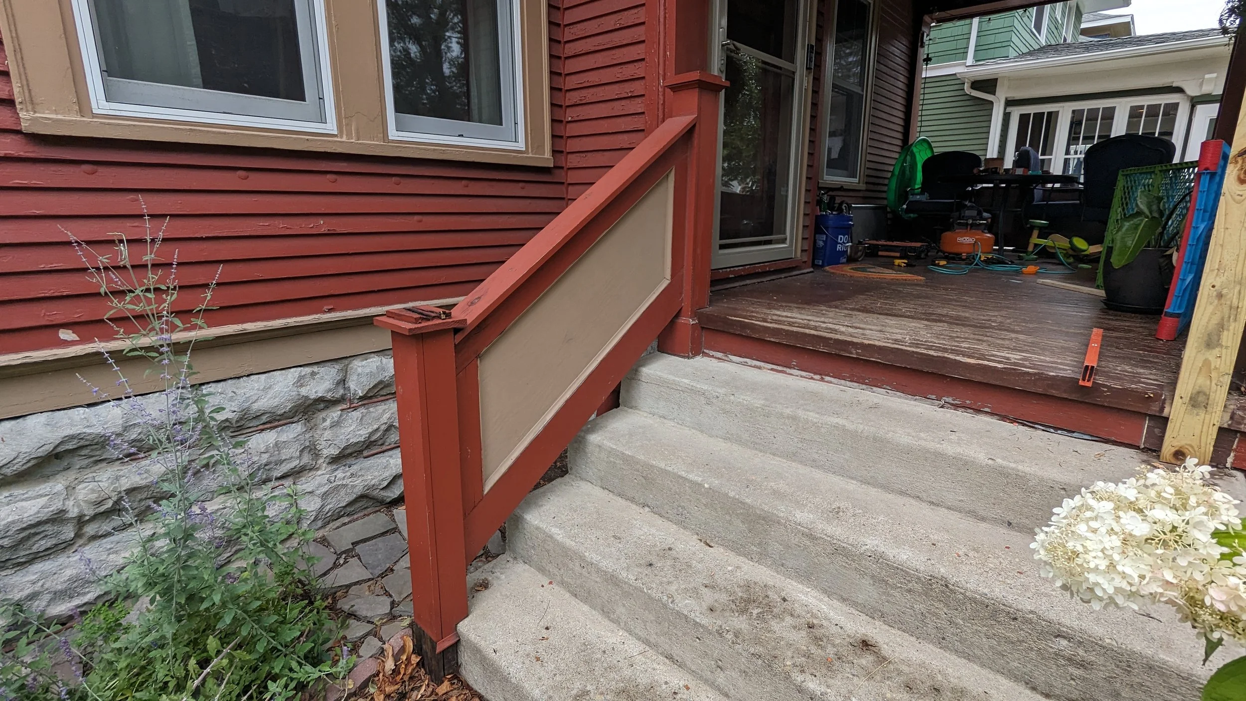 View of outdoor stairs leading up to a wooden porch at a house, with a red railing, concrete steps, and a storage area with various tools and items in the background.