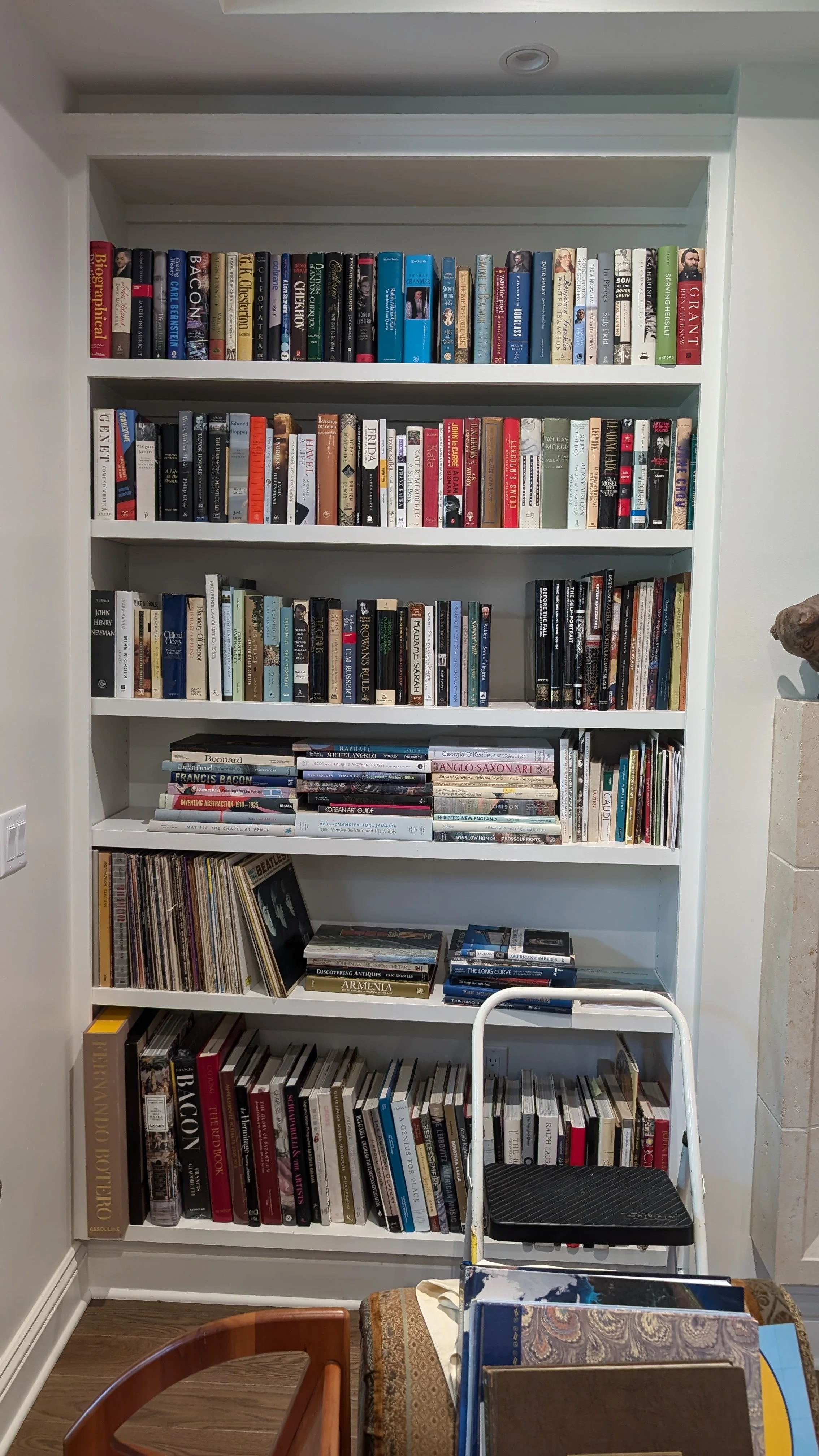 A white bookshelf filled with books in a room with hardwood floors, a small staircase, and a white wall. There are fourmain shelves and additional books below.