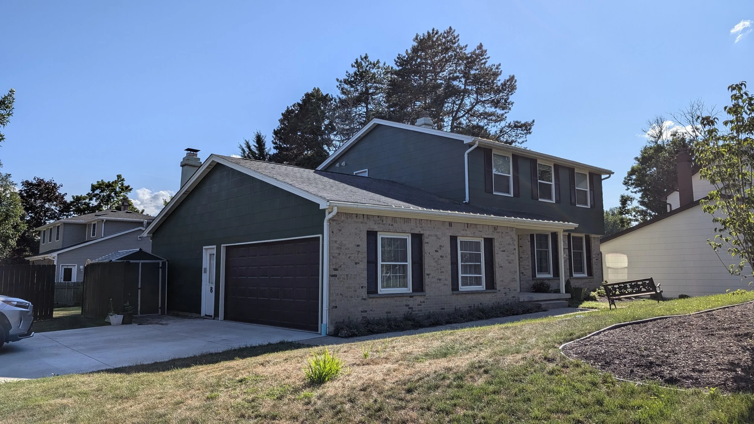 A two-story house with a brick and dark green siding exterior, a garage door, multiple windows with black shutters, a driveway, a small lawn with a bench, a tree in the background, and a bright blue sky.