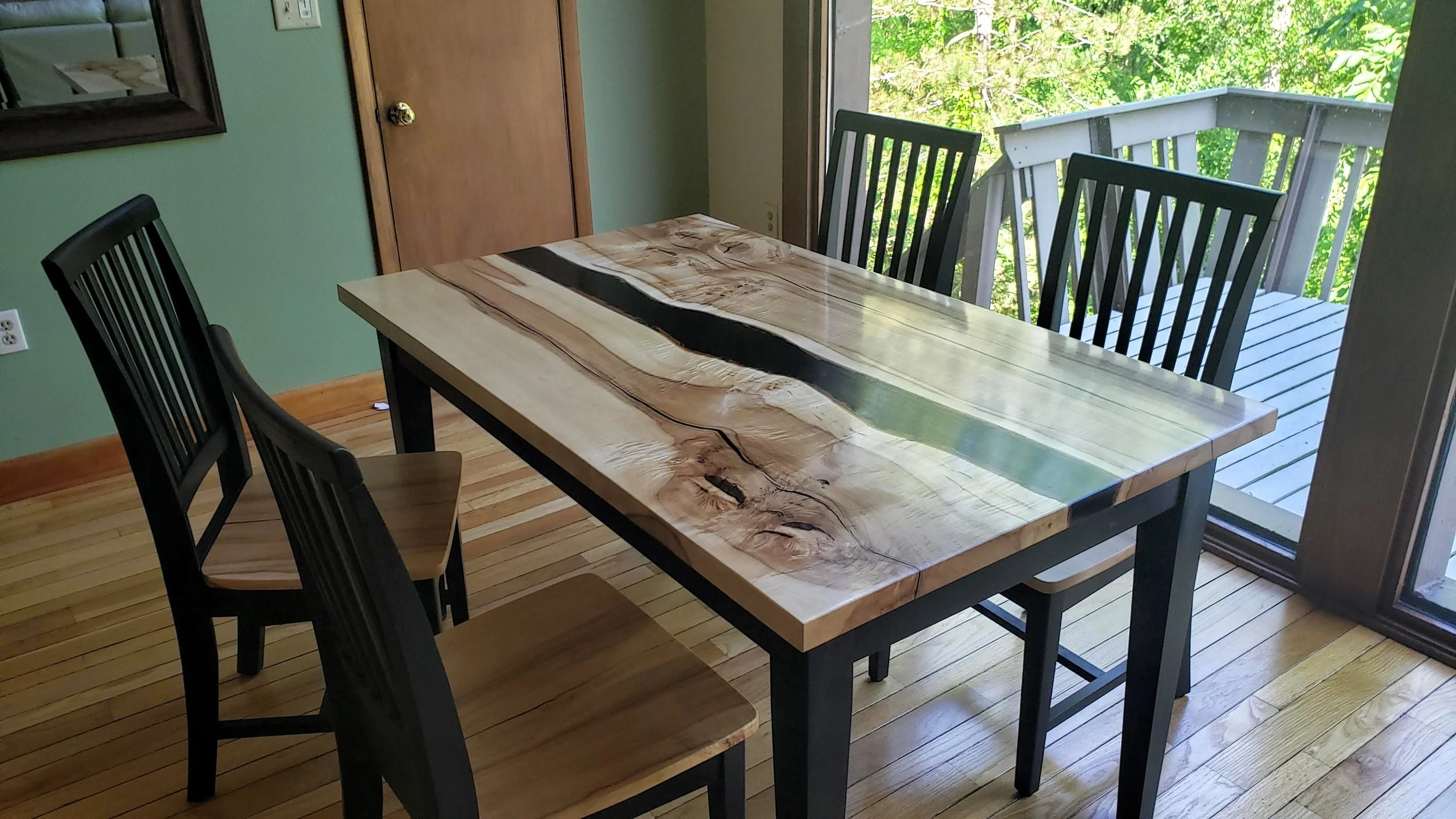 Wooden dining table with black legs and three black chairs on a hardwood floor, with a sliding glass door showing trees outside.