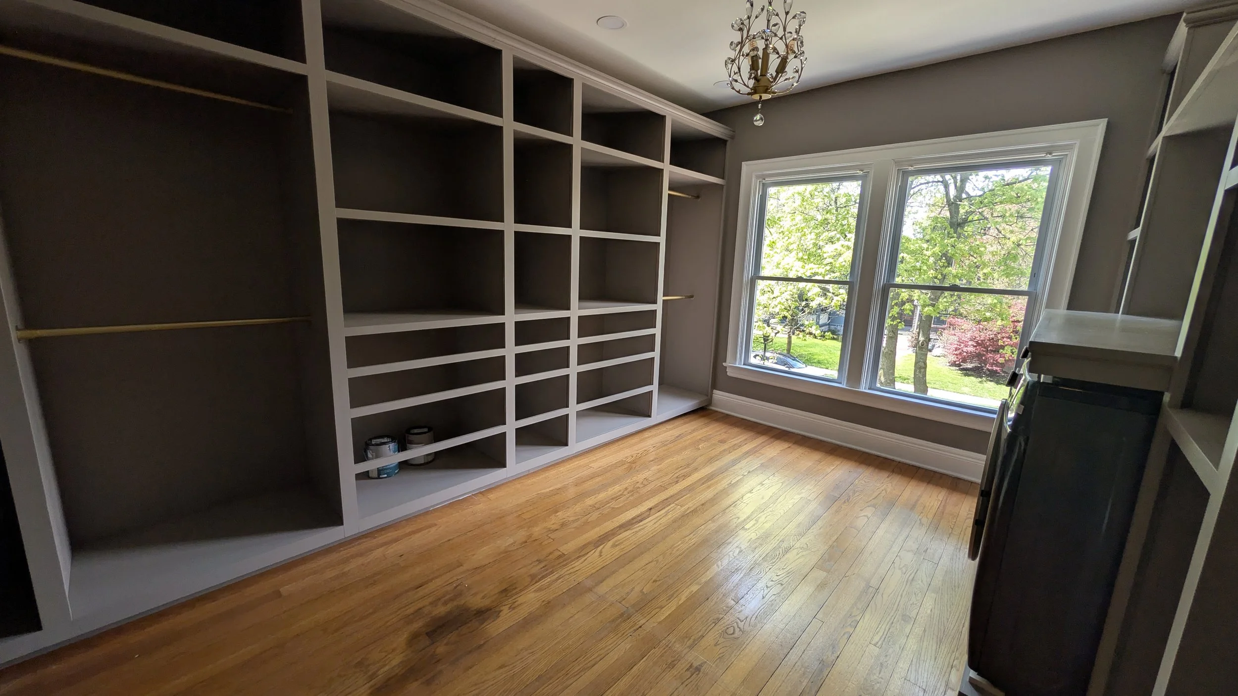 Empty built-in shelving units in a room with large windows showing a tree outside, hardwood flooring, and a chandelier ceiling light.