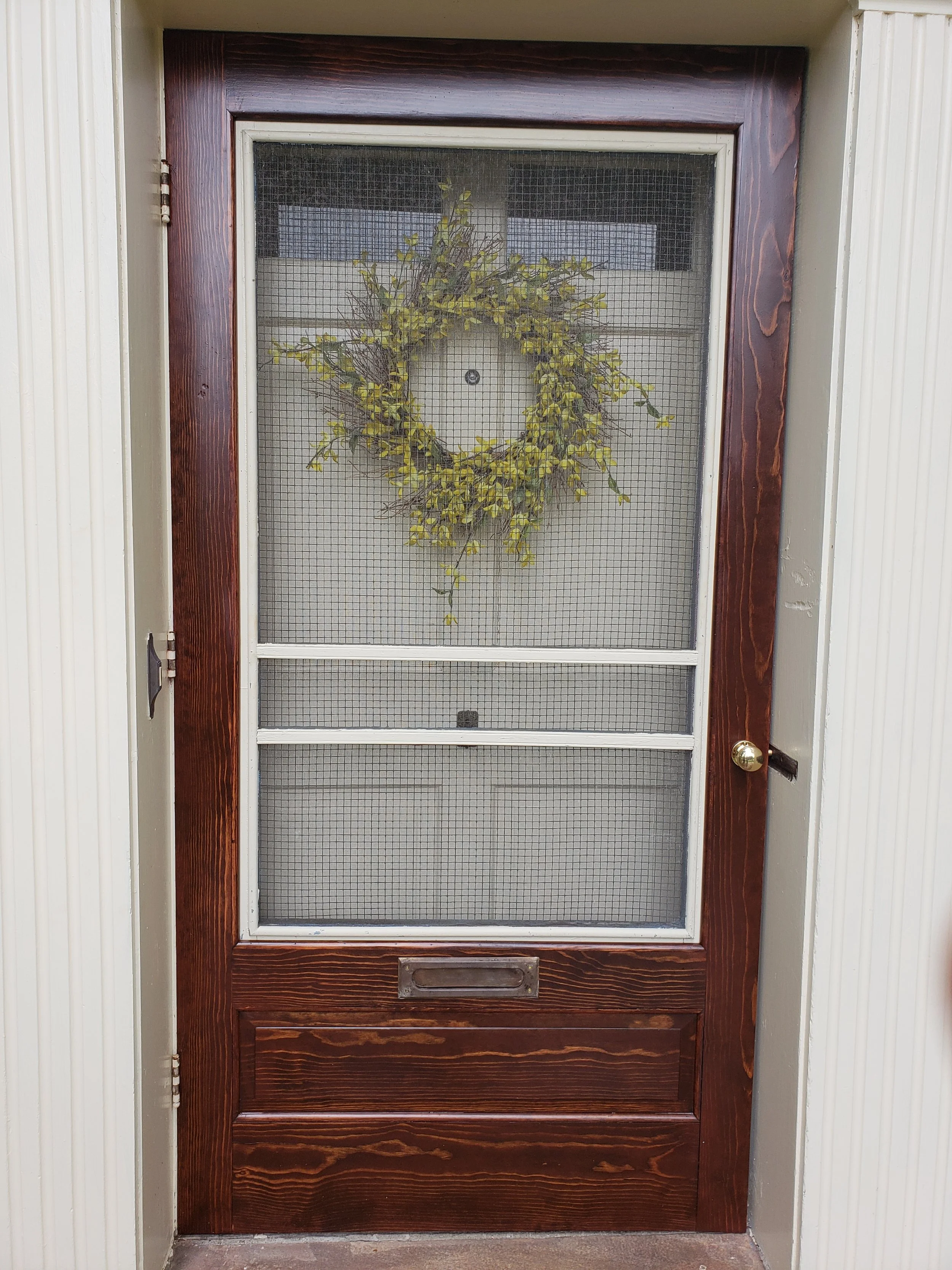 A wooden door with a metal screen in the upper and middle sections, featuring a wreath made of green and brown foliage, and a small mail slot at the bottom.