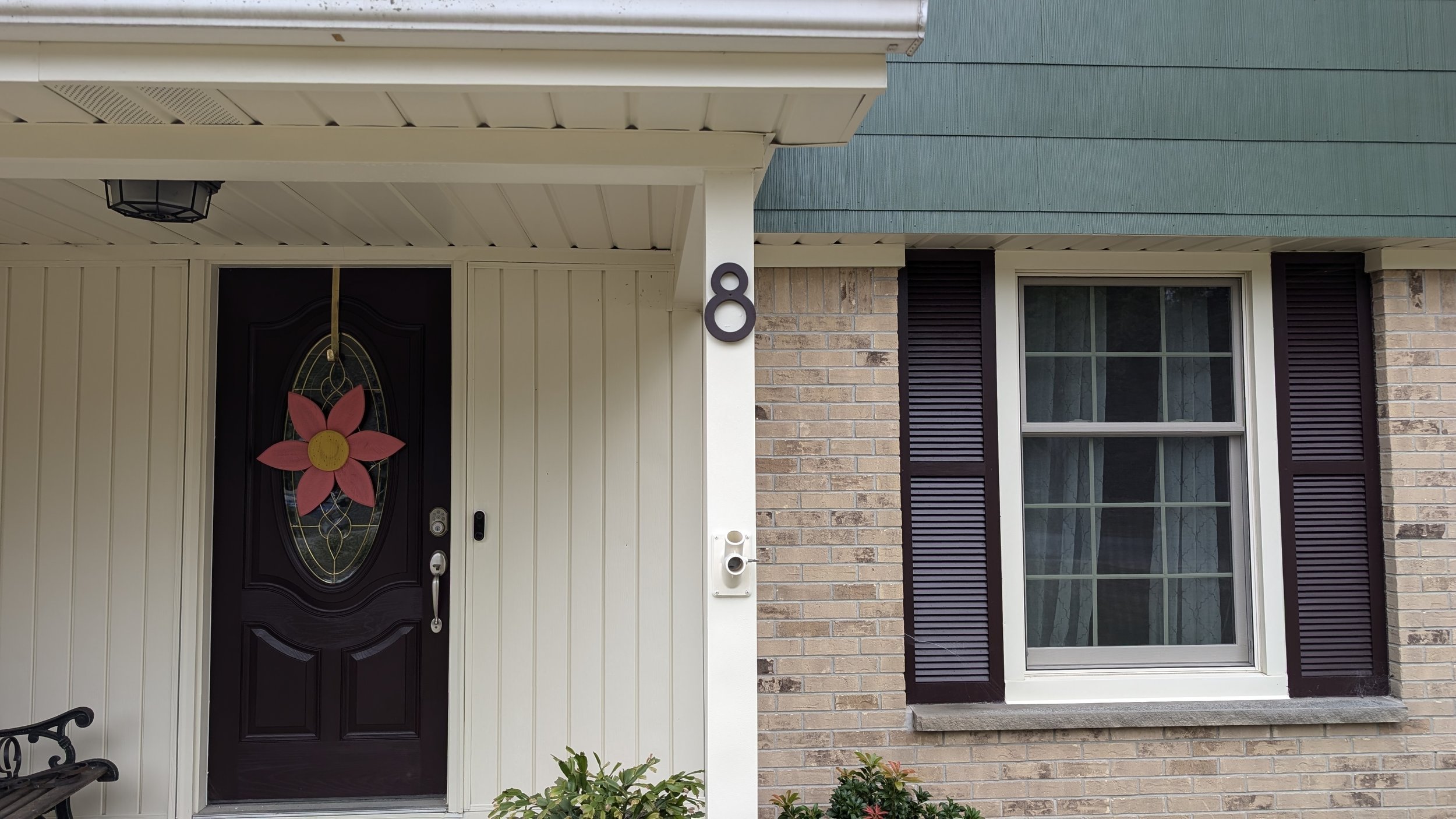 Close-up of house front door with decorative flower wreath, house number 8 on siding, brick wall, and window above with black shutters.