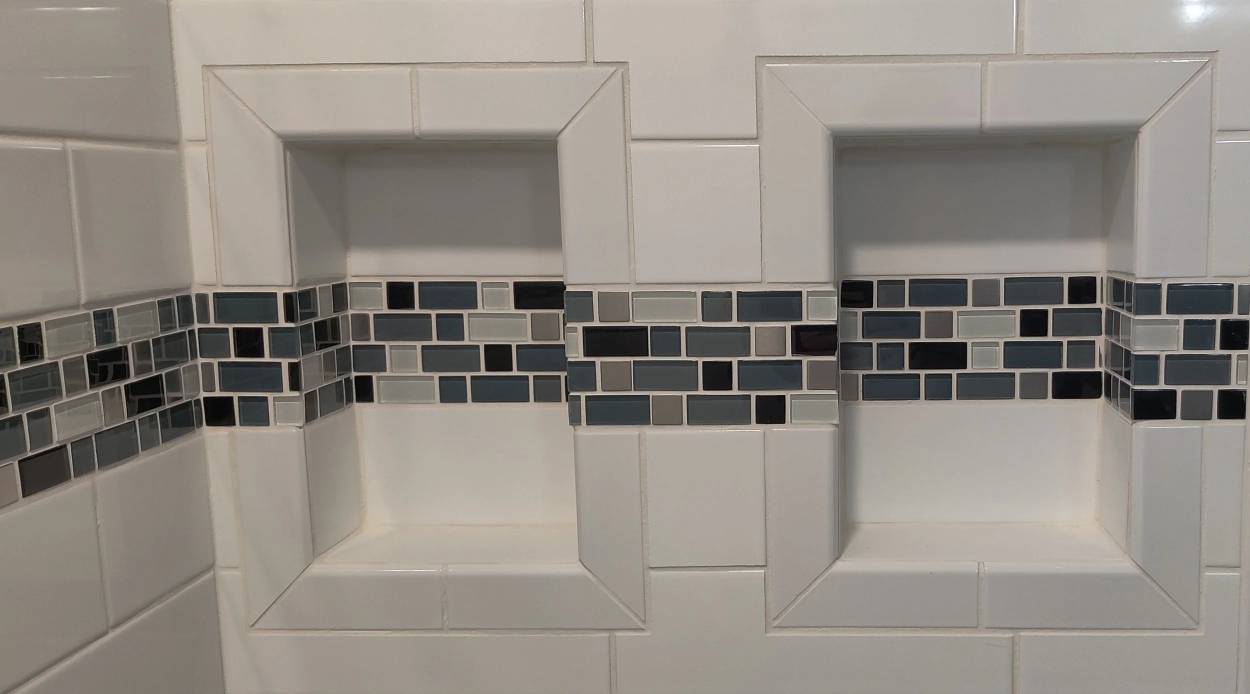Close-up of a tiled wall with two small recessed shelves outlined by a tiled border. The lower part of the wall has small, rectangular mosaic tiles in shades of gray, black, and white.