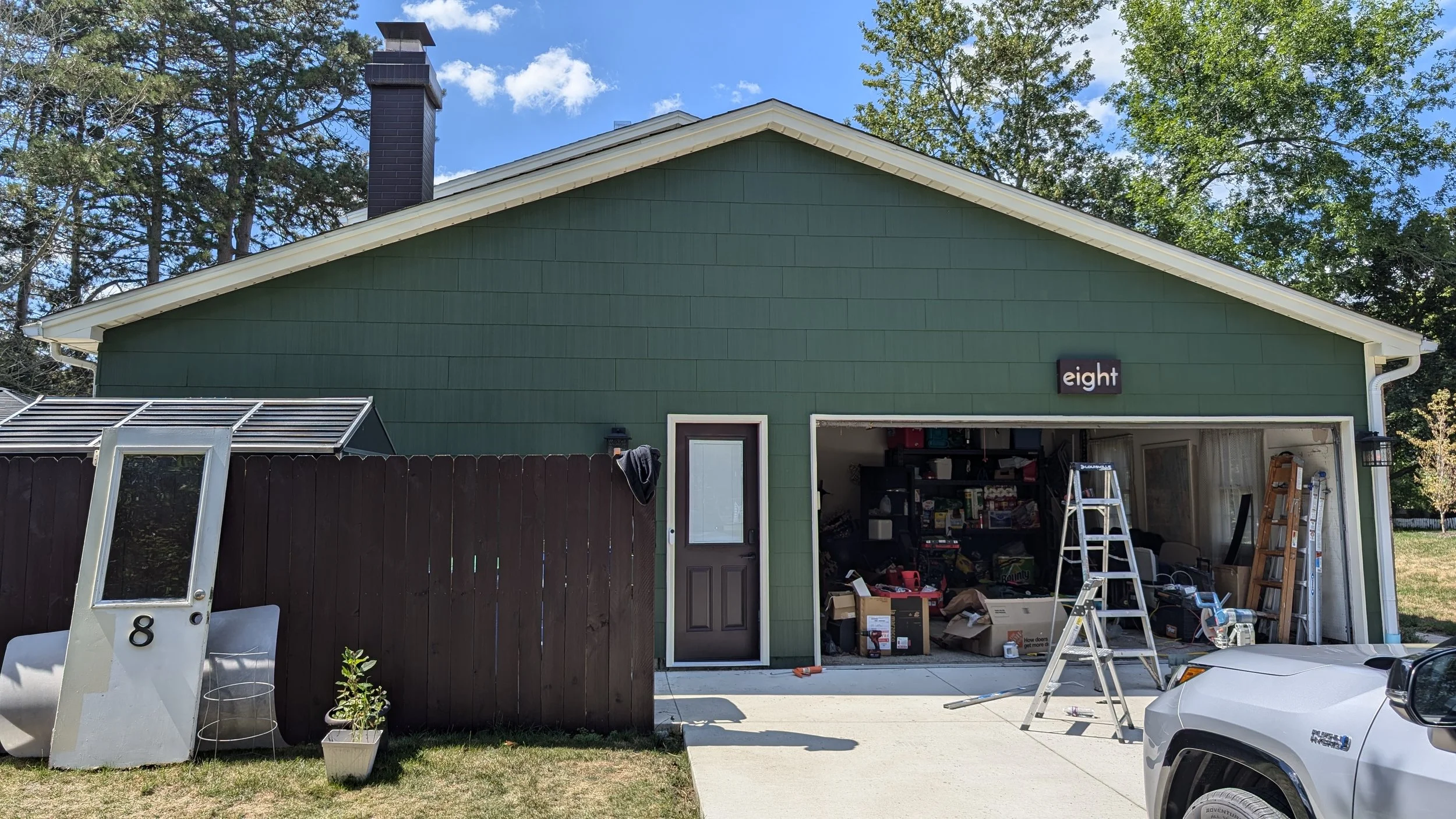Backyard scene showing a green house with a garage, which is open and cluttered with tools, boxes, and ladders. A white mailbox with the number 8 is in the foreground, along with potted plants, a small greenhouse, and part of a white car. The sky is 