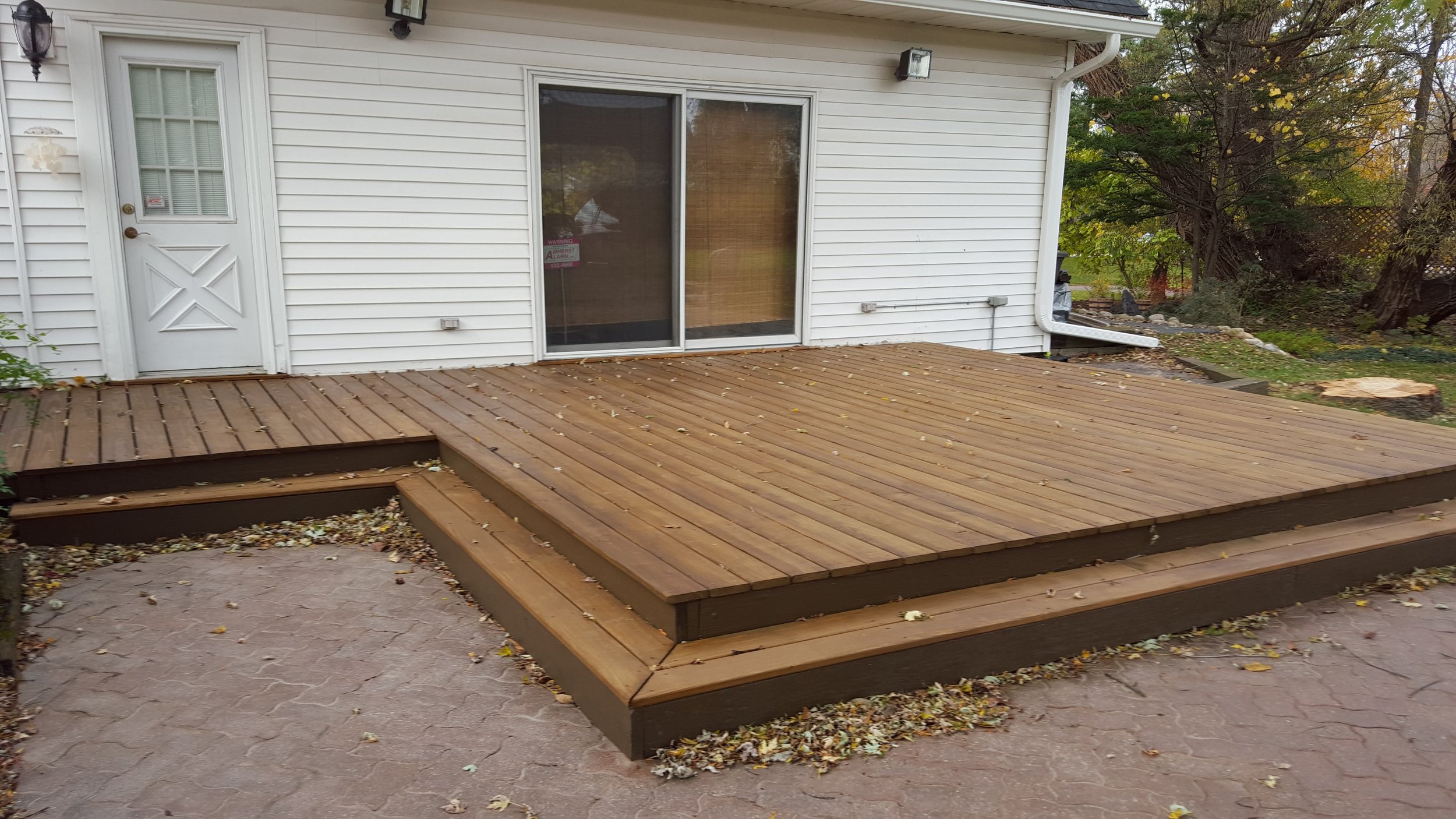 A wooden deck attached to the back of a house with white siding, sliding glass door, and surrounding trees with autumn leaves.