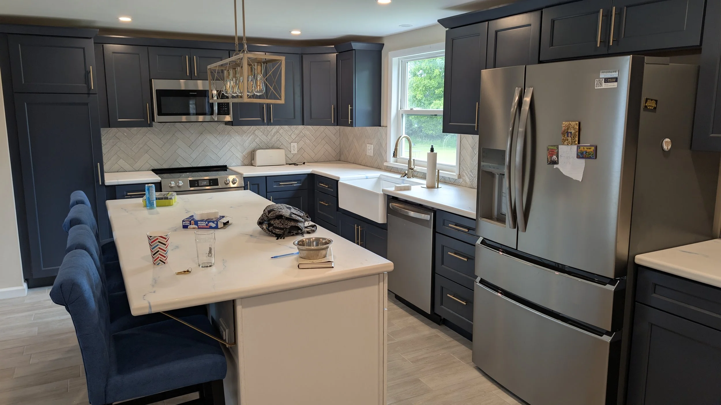 Modern kitchen with dark blue cabinets, a white island, stainless steel appliances, and a window overlooking greenery.