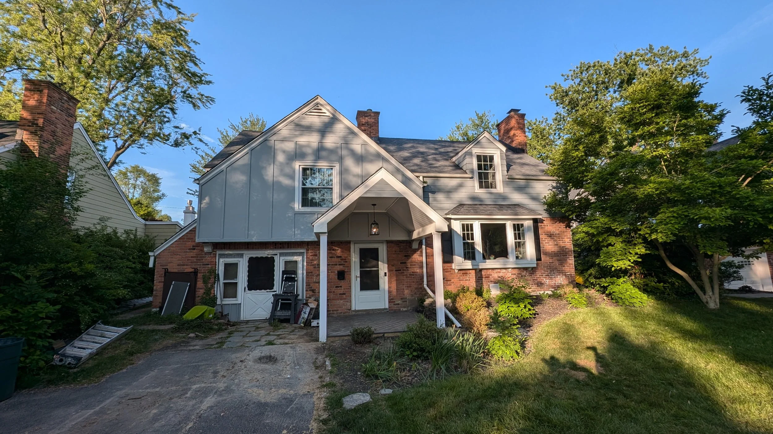 A two-story house with a gabled roof, brick and light-colored siding exterior, large windows, and a small front porch with a hanging lantern. The yard has trees, shrubs, a paved pathway, and some clutter near the house