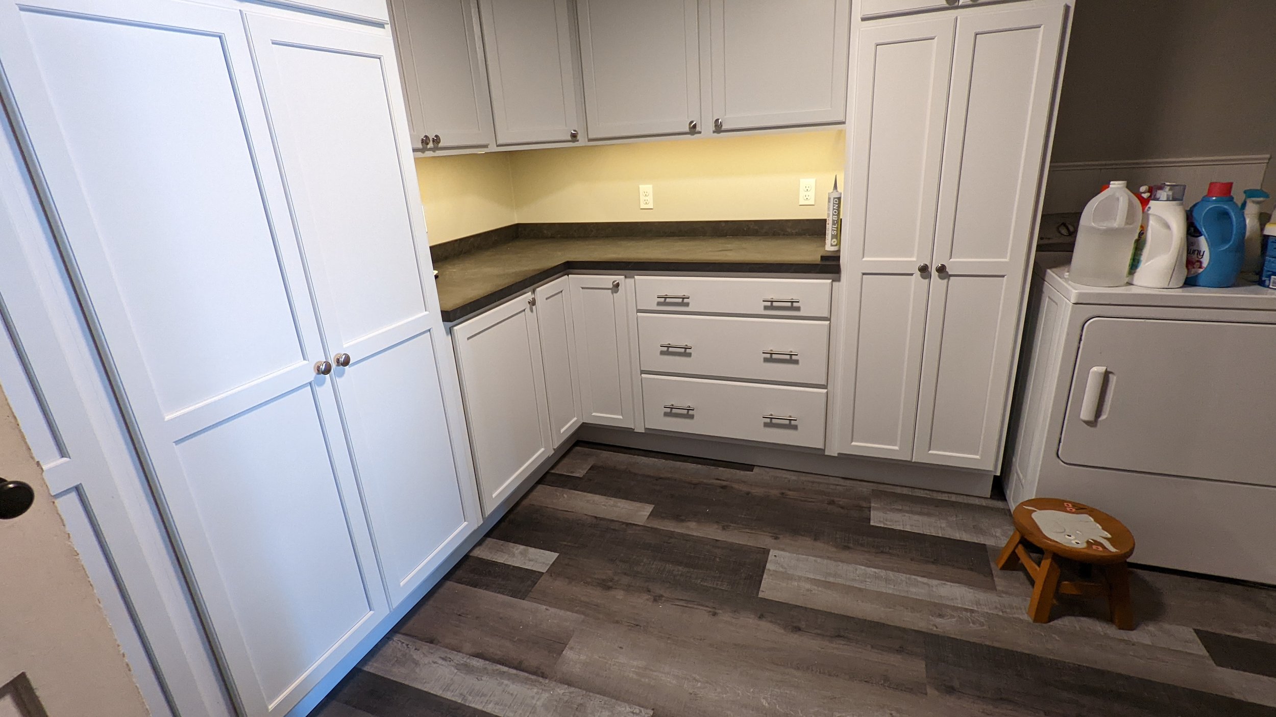 White kitchen cabinets with black handles, a countertop with a tube of silicone sealant, laundry detergent bottles on a washing machine, and a small wooden stool with cartoon character print.