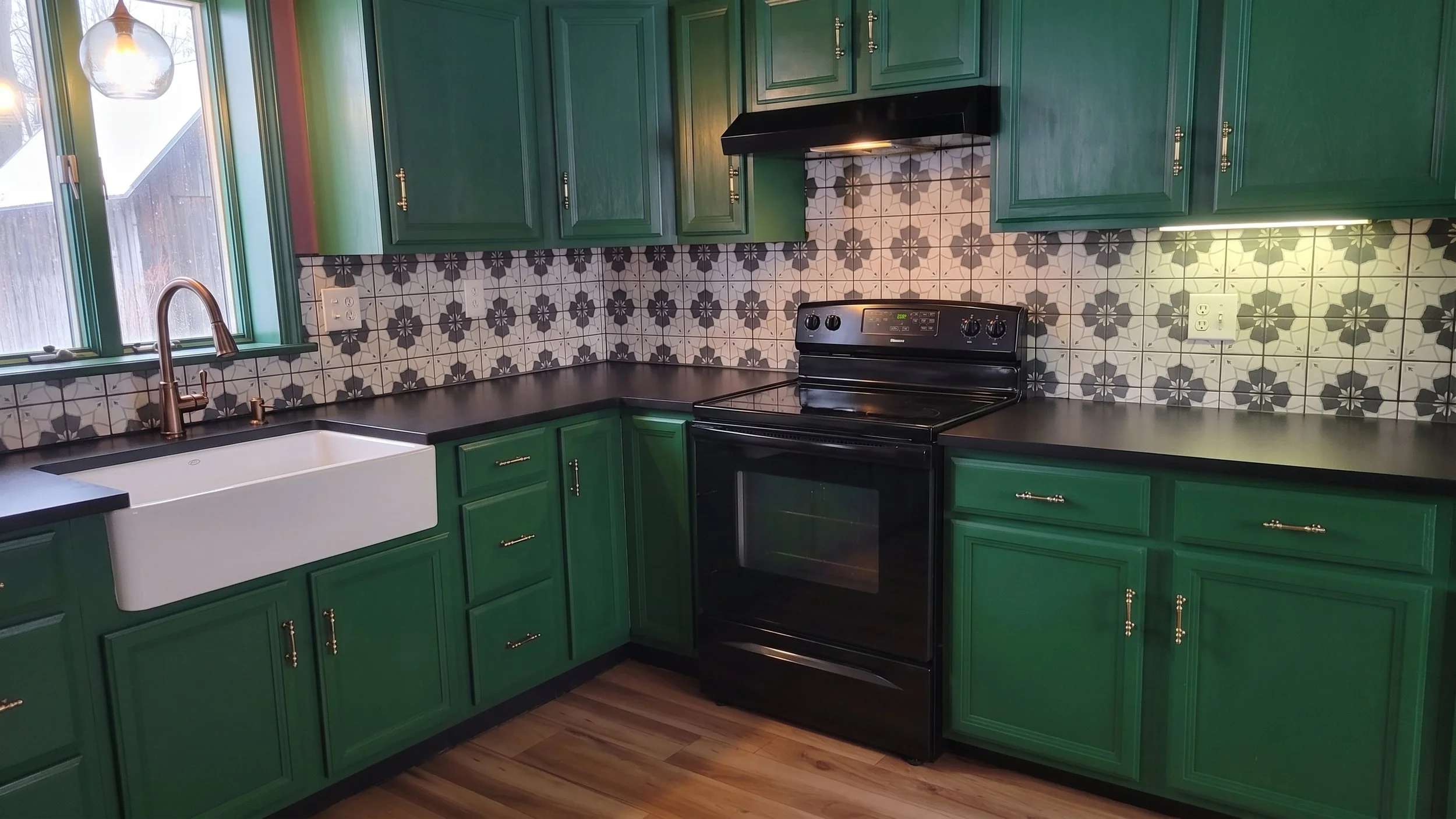 A kitchen with green cabinets, black countertops, a white farmhouse sink, black stove, patterned tile backsplash, window, and hardwood floors.