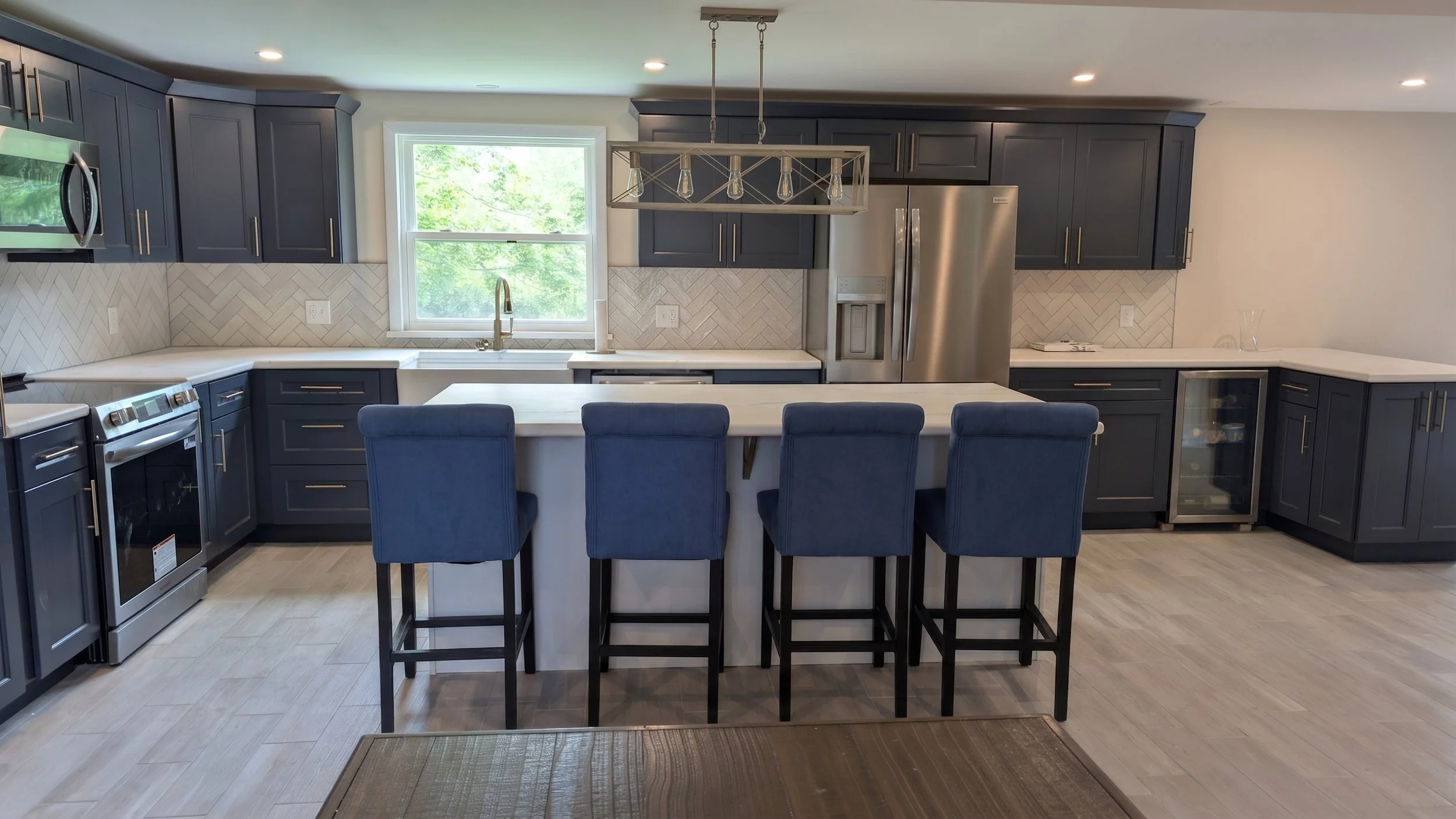 Modern kitchen with dark blue cabinets, white countertops, and stainless steel appliances, including a refrigerator, microwave, and oven. A central island with seating for four has four blue chairs. There is a window above the sink with green trees o